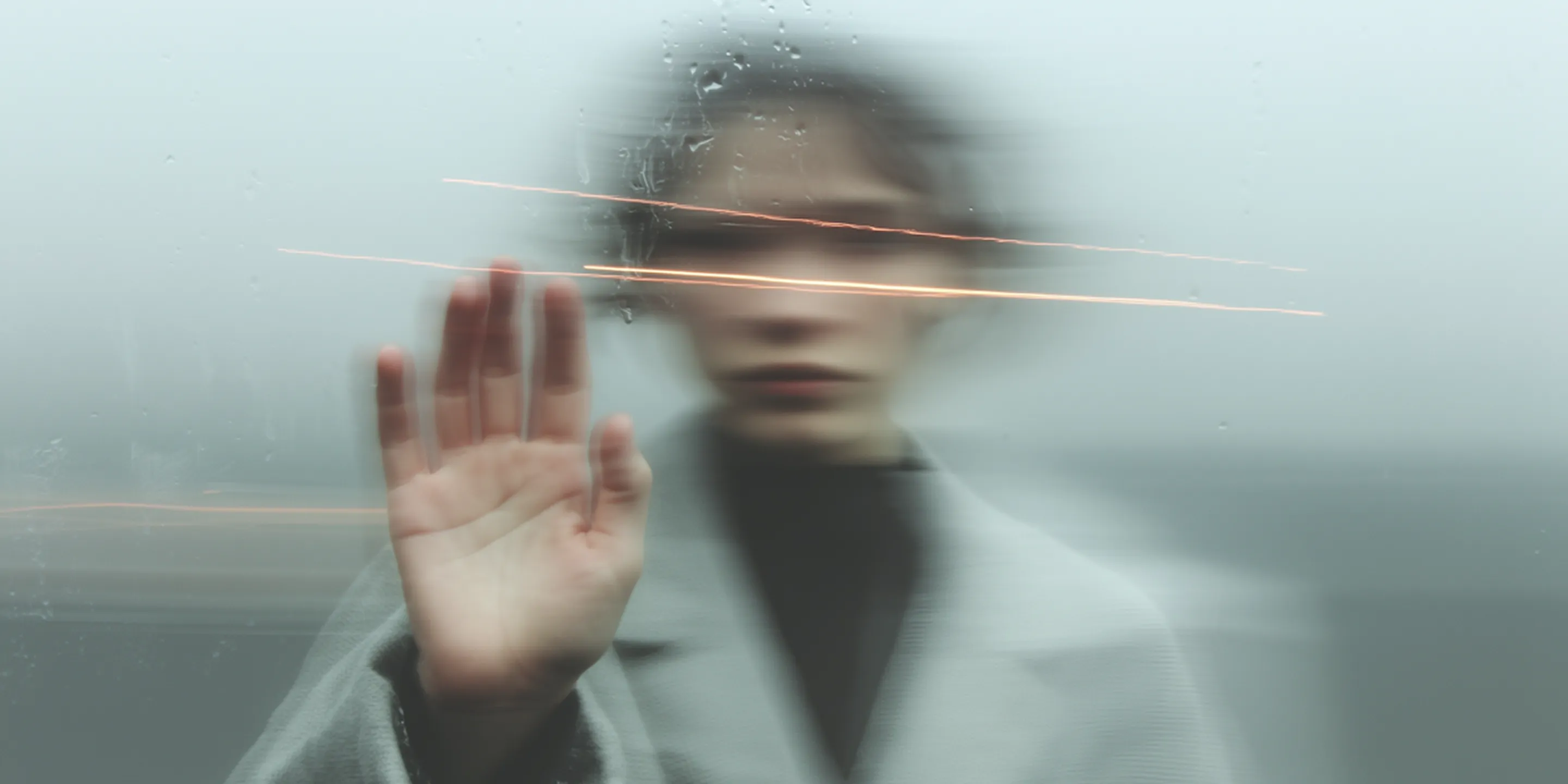 Blurred person pressing their hand against wet glass with light streaks across face.