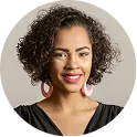 Smiling young woman with curly hair wearing a black top and red earrings against a beige background.