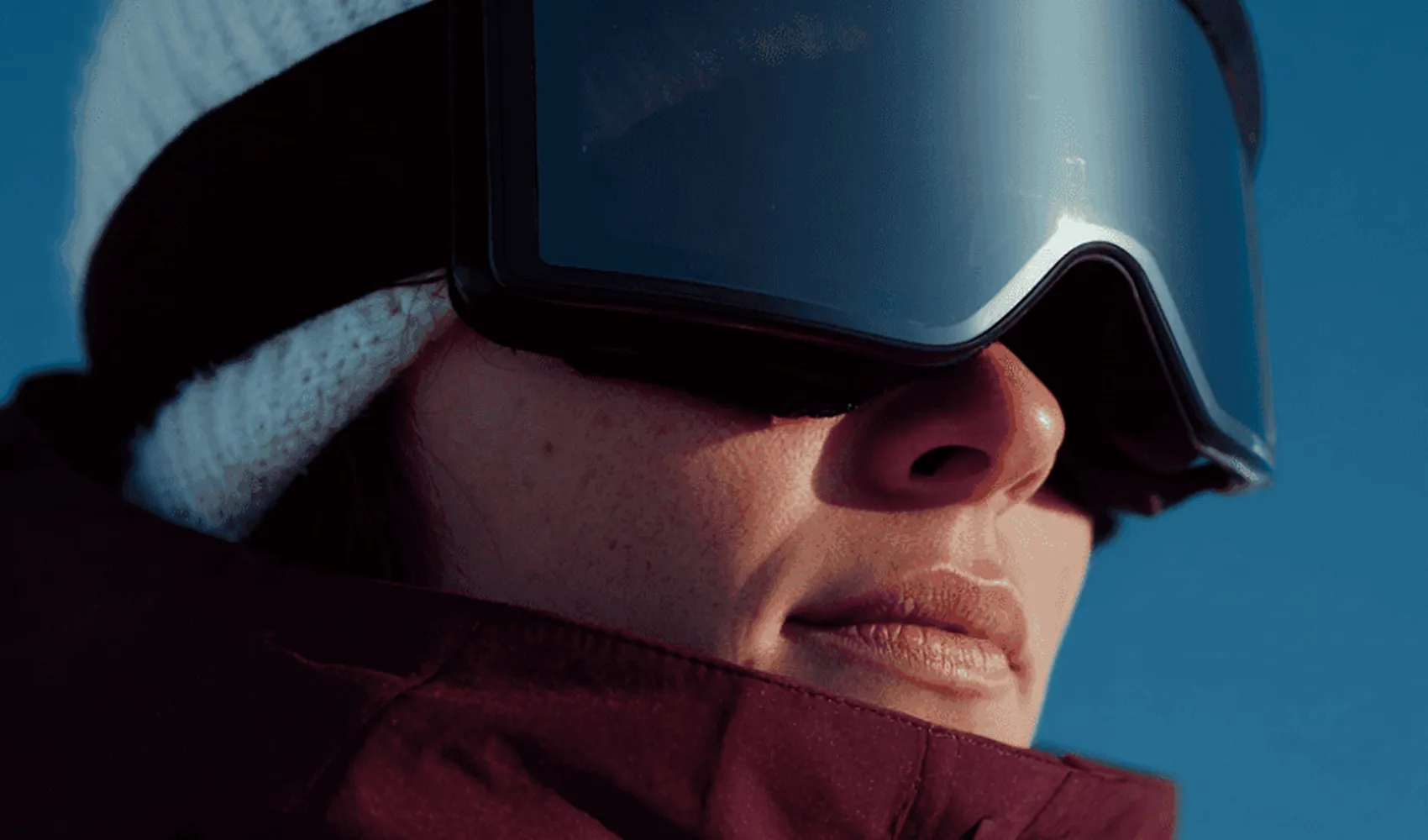 Close-up of person wearing a white knit hat and large ski goggles against a blue sky.