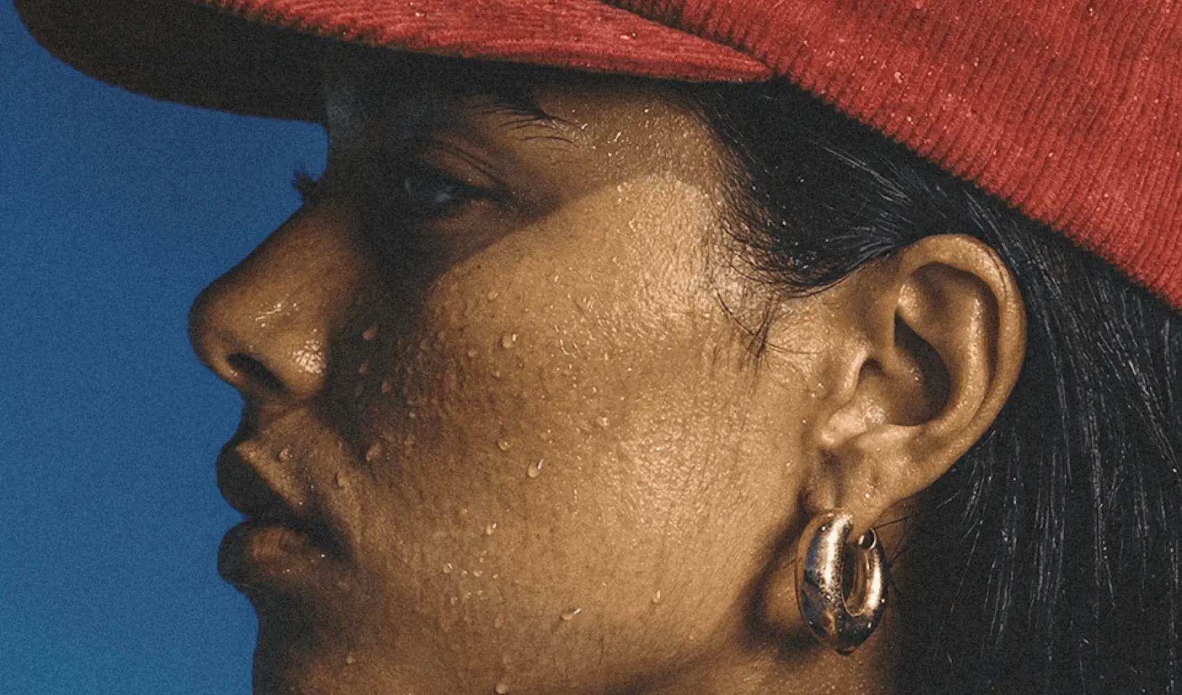 Close-up side profile of a person wearing a red hat, gold hoop earring, and sweat on skin.