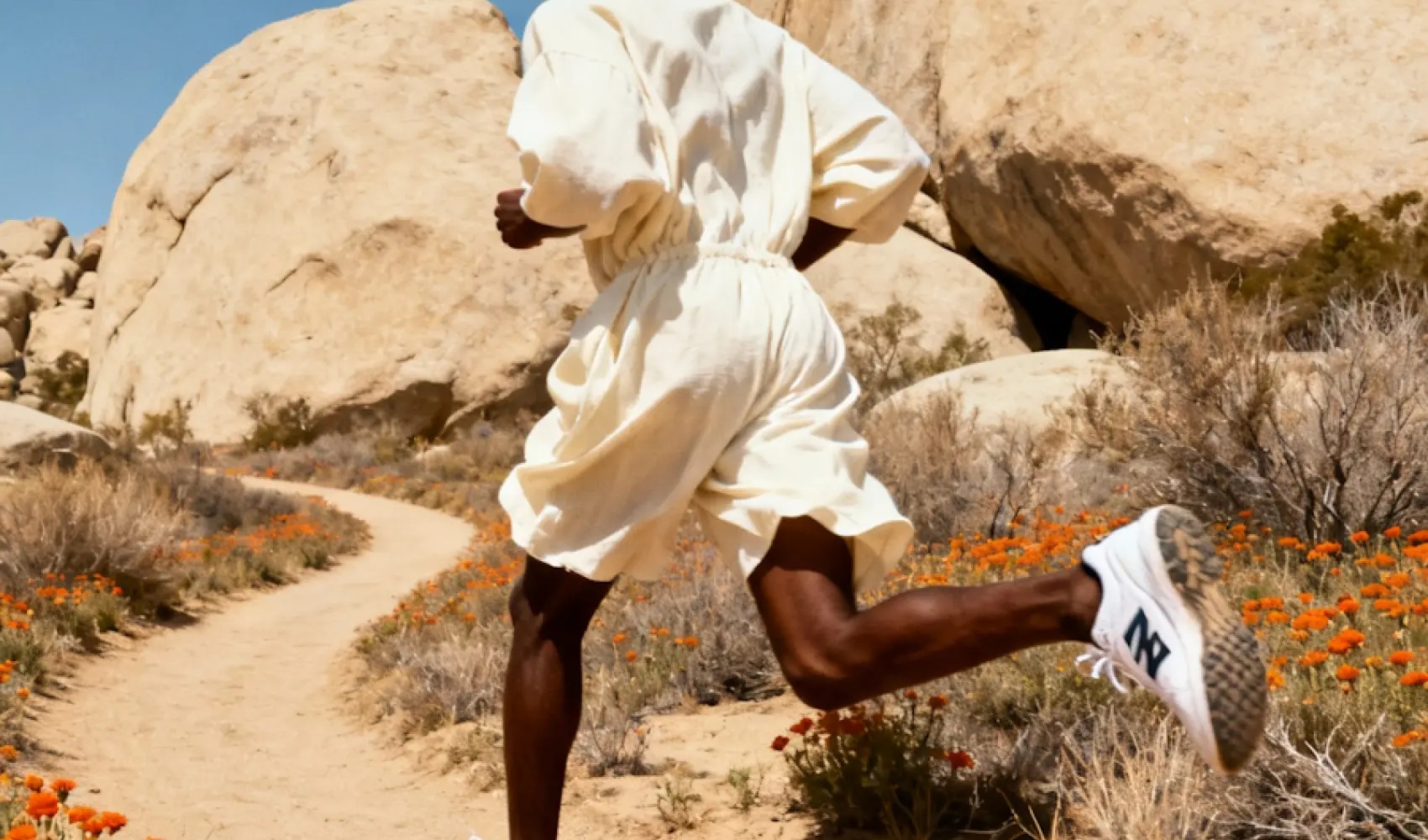 Person in a flowing beige outfit running on a rocky desert path with orange flowers.