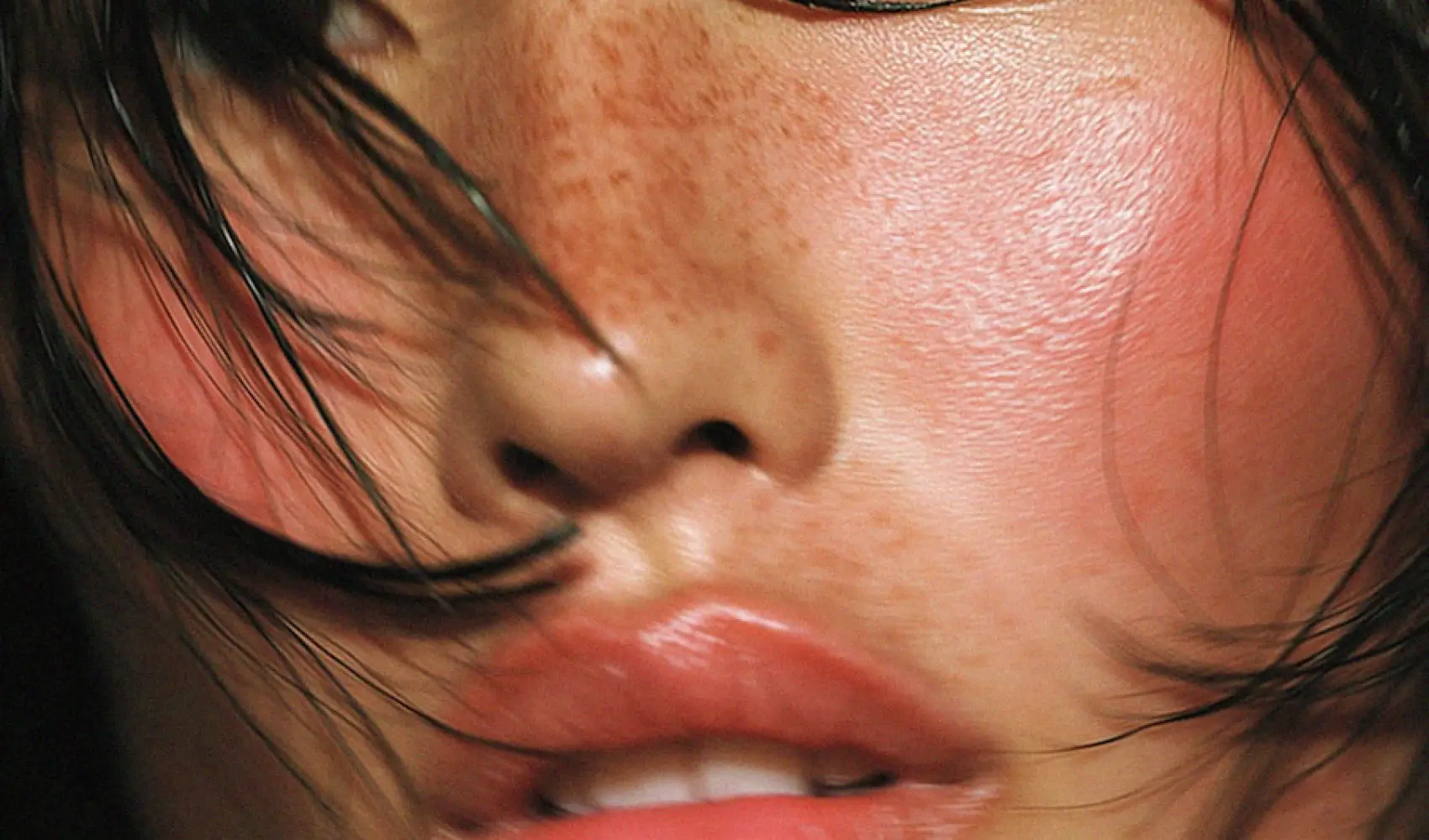 Close-up of a person's freckled face with red cheeks, lips, and strands of dark hair.
