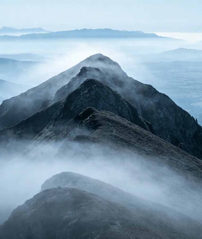 Layered mountain peaks with mist swirling around the dark rocky ridges under a pale sky.
