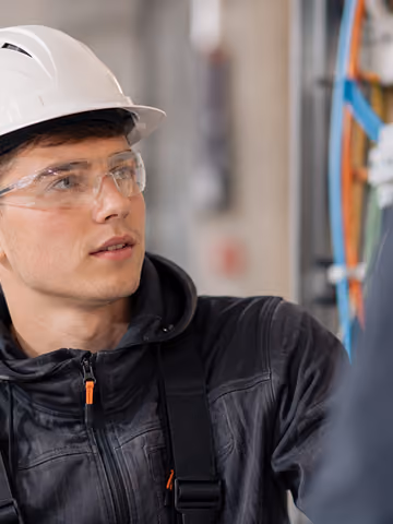 Junger Elektriker mit Helm und Schutzbrille prüft Kabel in einem technischen Raum.