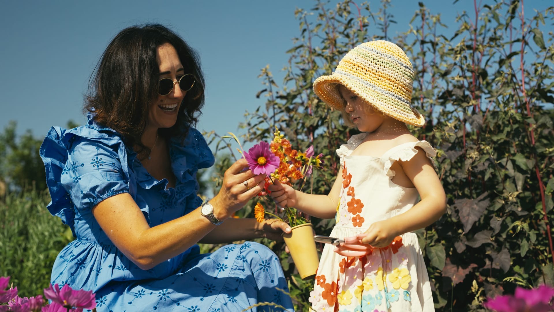 A woman and a small child in a sun hat picking bright flowers together in the field