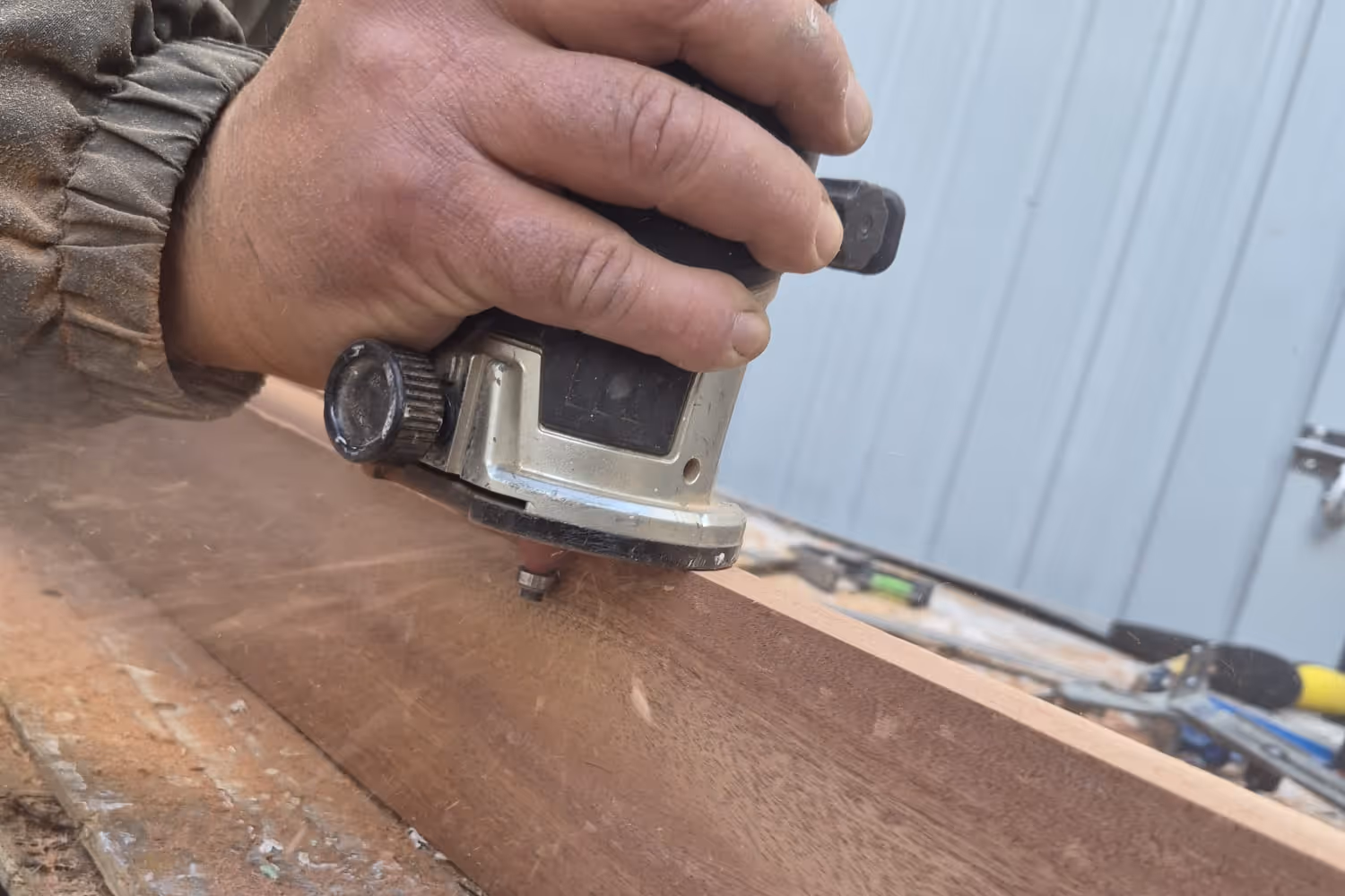 Hand holding a wood router smoothing the edge of a wooden plank on a workbench.