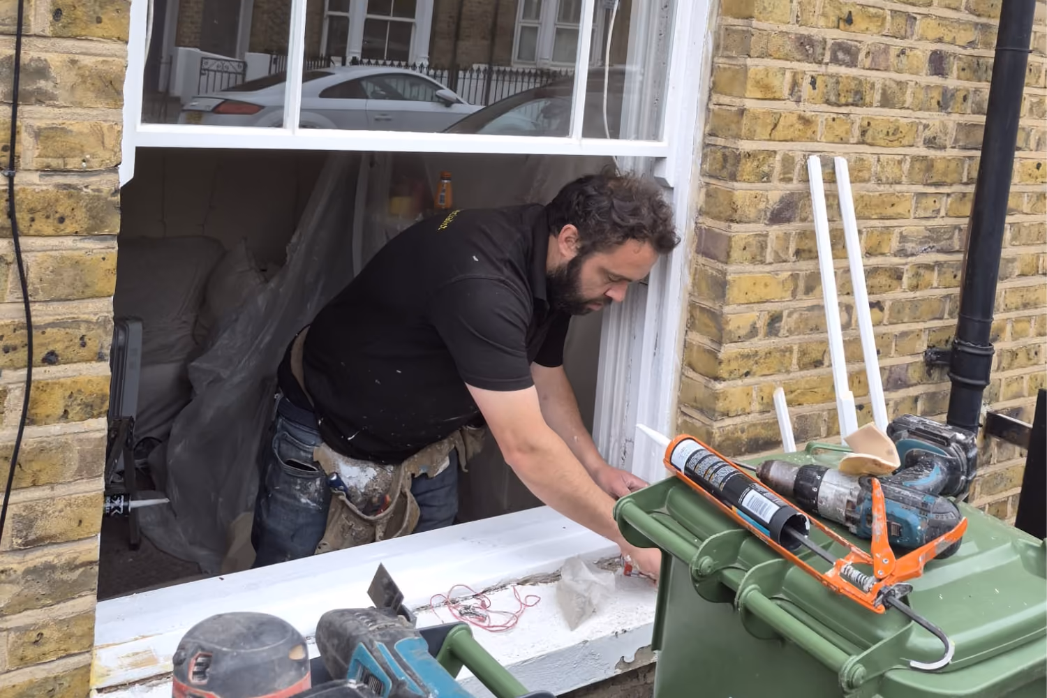 Worker repairing a window frame of a brick house using tools and sealant during renovation.