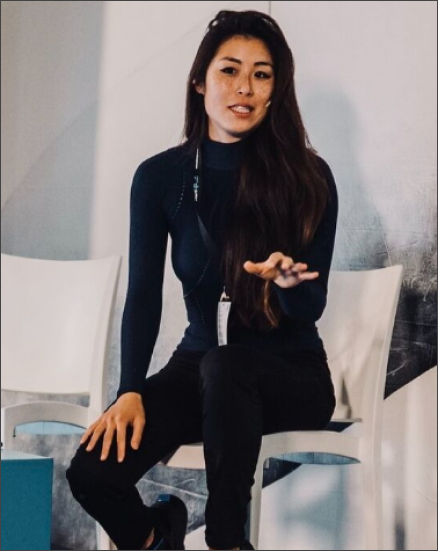Woman with long hair in black outfit sitting on a chair, gesturing with her hand while speaking.
