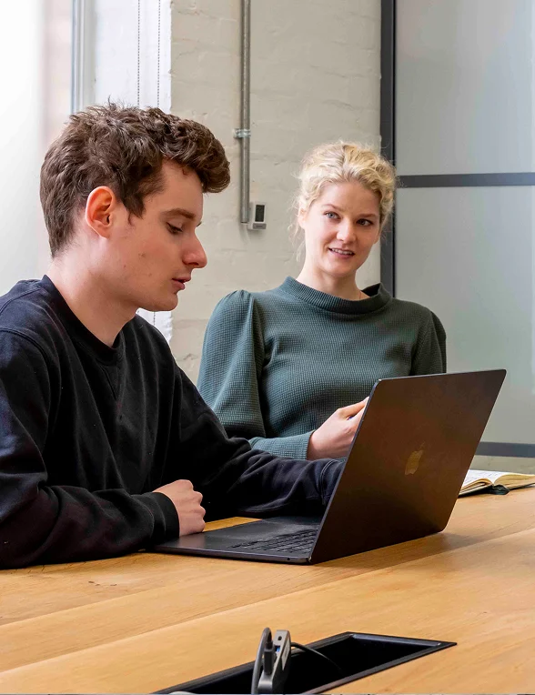 Two people sitting at a wooden table, one using a laptop, engaged in conversation in a modern office.