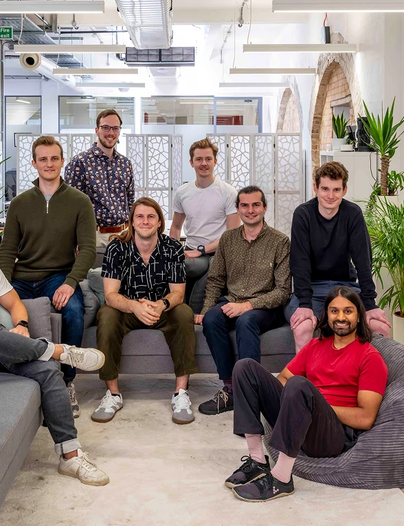 Group of eight casually dressed men posing together in a bright, modern office lounge area.