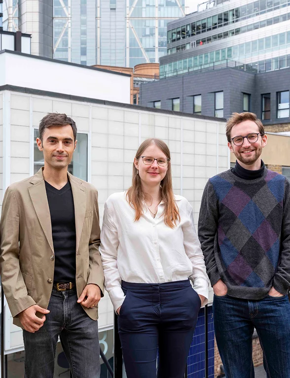 Three young professionals standing outdoors with modern buildings in the background, smiling at the camera.
