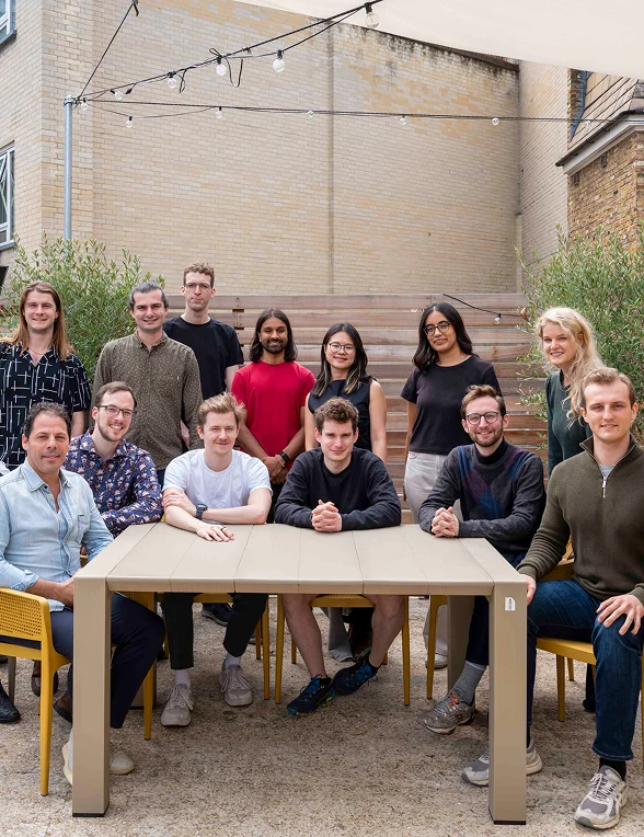 Group of thirteen diverse people seated and standing around a table outdoors against a brick wall.