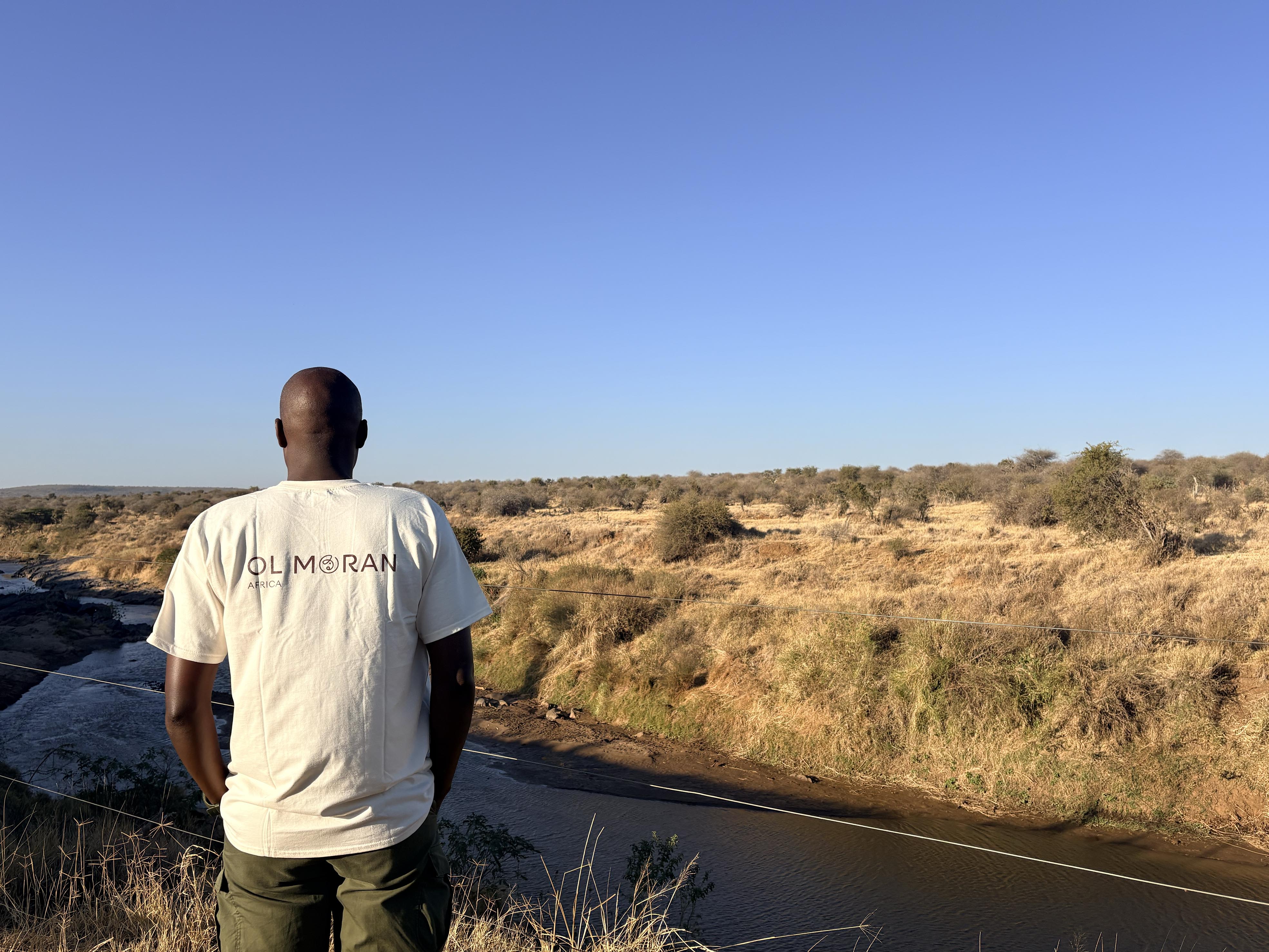 A man wearing a white Ol Moran Africa t-shirt stands with his back to the camera, overlooking a river and dry grassy plains under a clear blue sky.