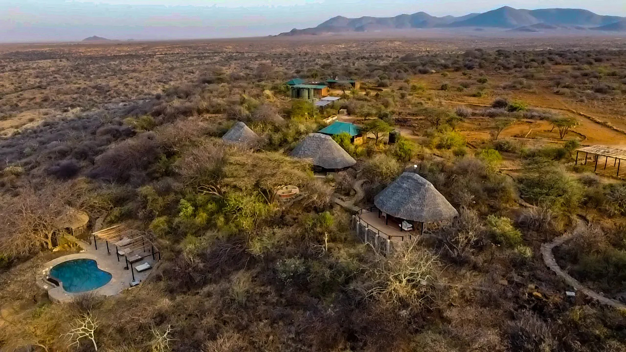 Aerial view of a rustic lodge with several thatched-roof huts and a swimming pool surrounded by dry savanna vegetation and distant mountains under a clear sky.