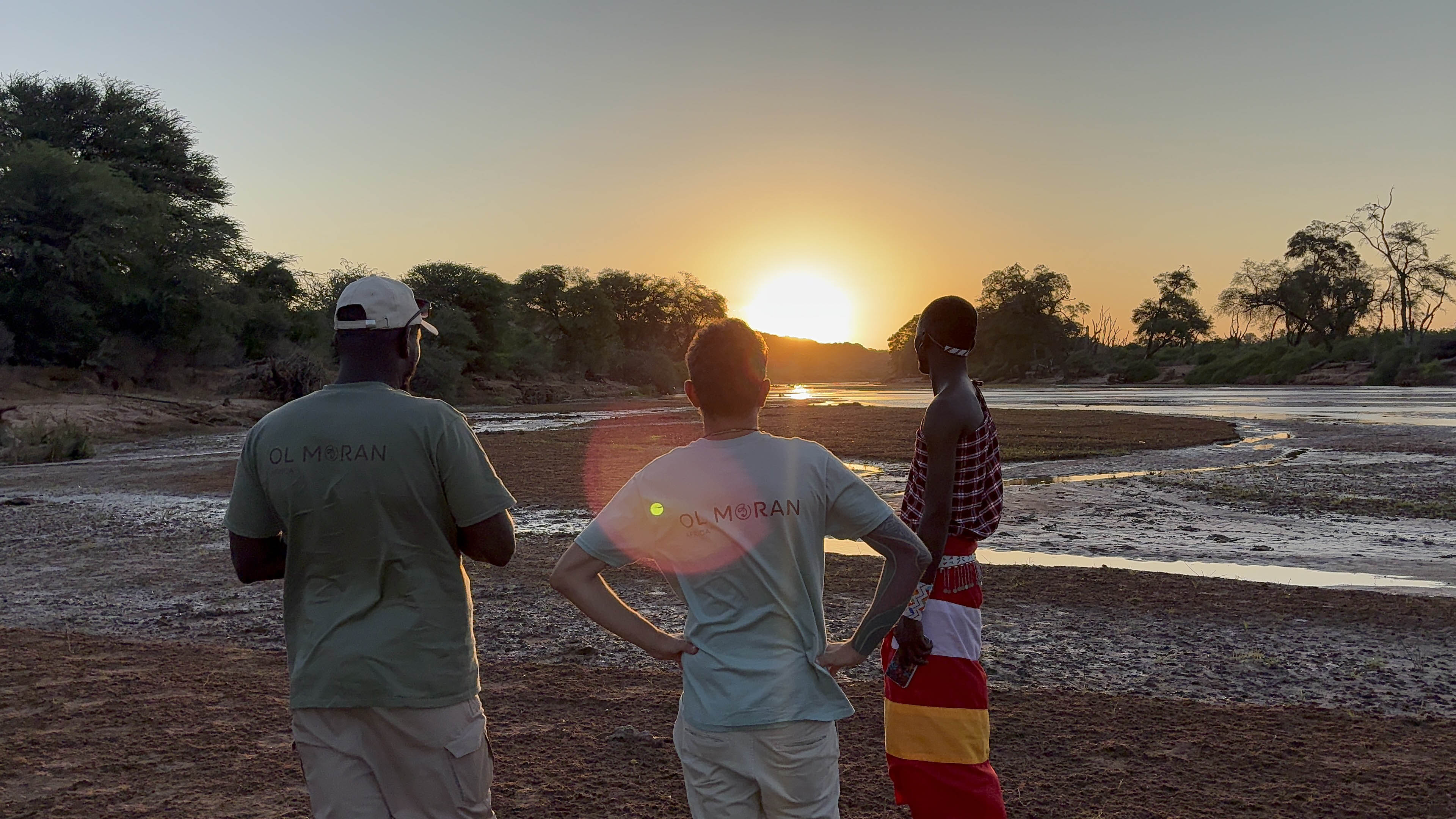 Three people stand on a riverbank at sunset, facing the horizon. Two wear green and light blue t-shirts with 'OL MORAN' on the back, while the third wears a red, yellow, and white traditional patterned wrap. Trees and water channels are visible around them.