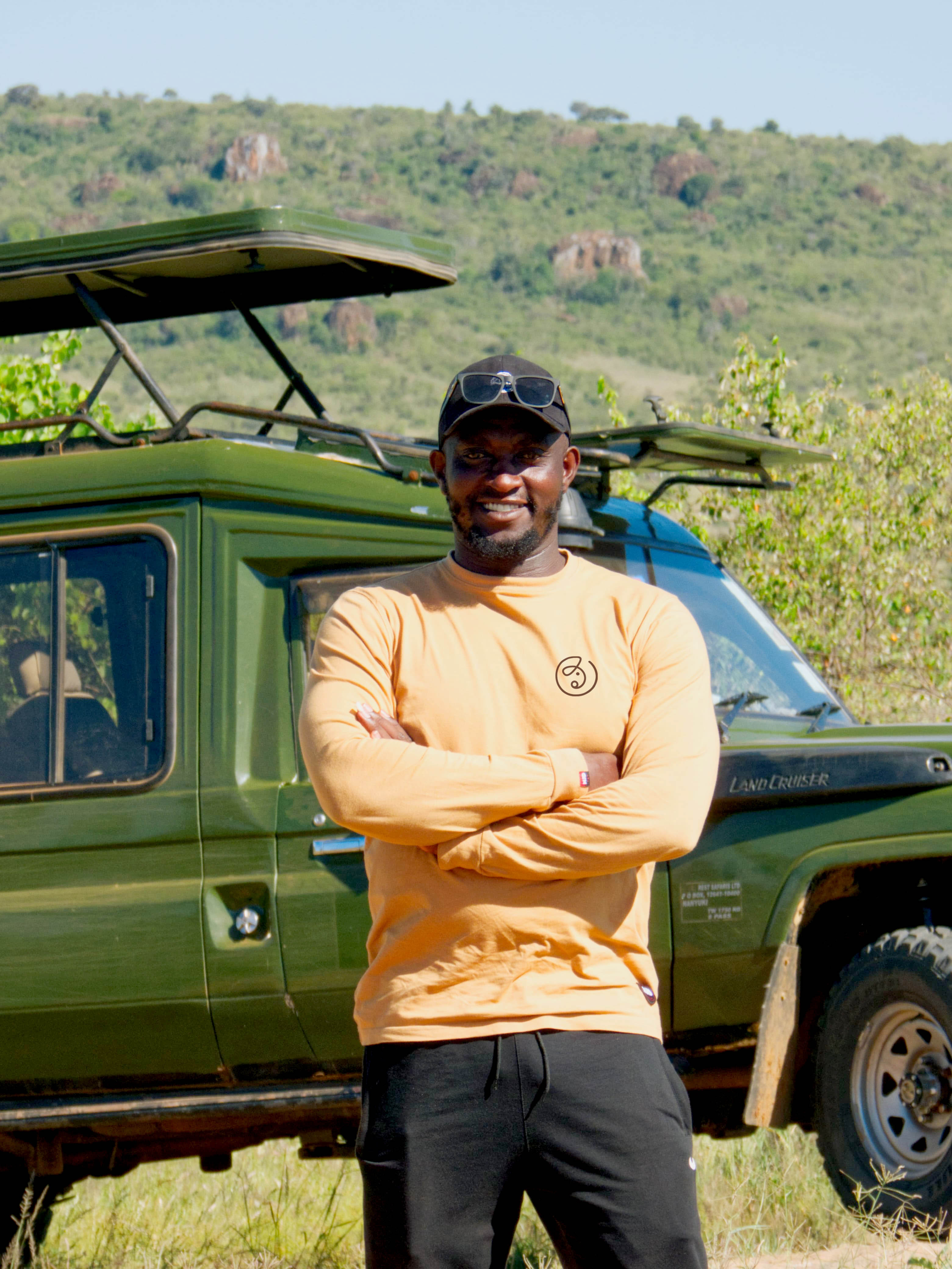 Smiling man wearing a black cap and orange long sleeve shirt with folded arms standing in front of a green Land Cruiser off-road vehicle in a grassy area with hills in the background.