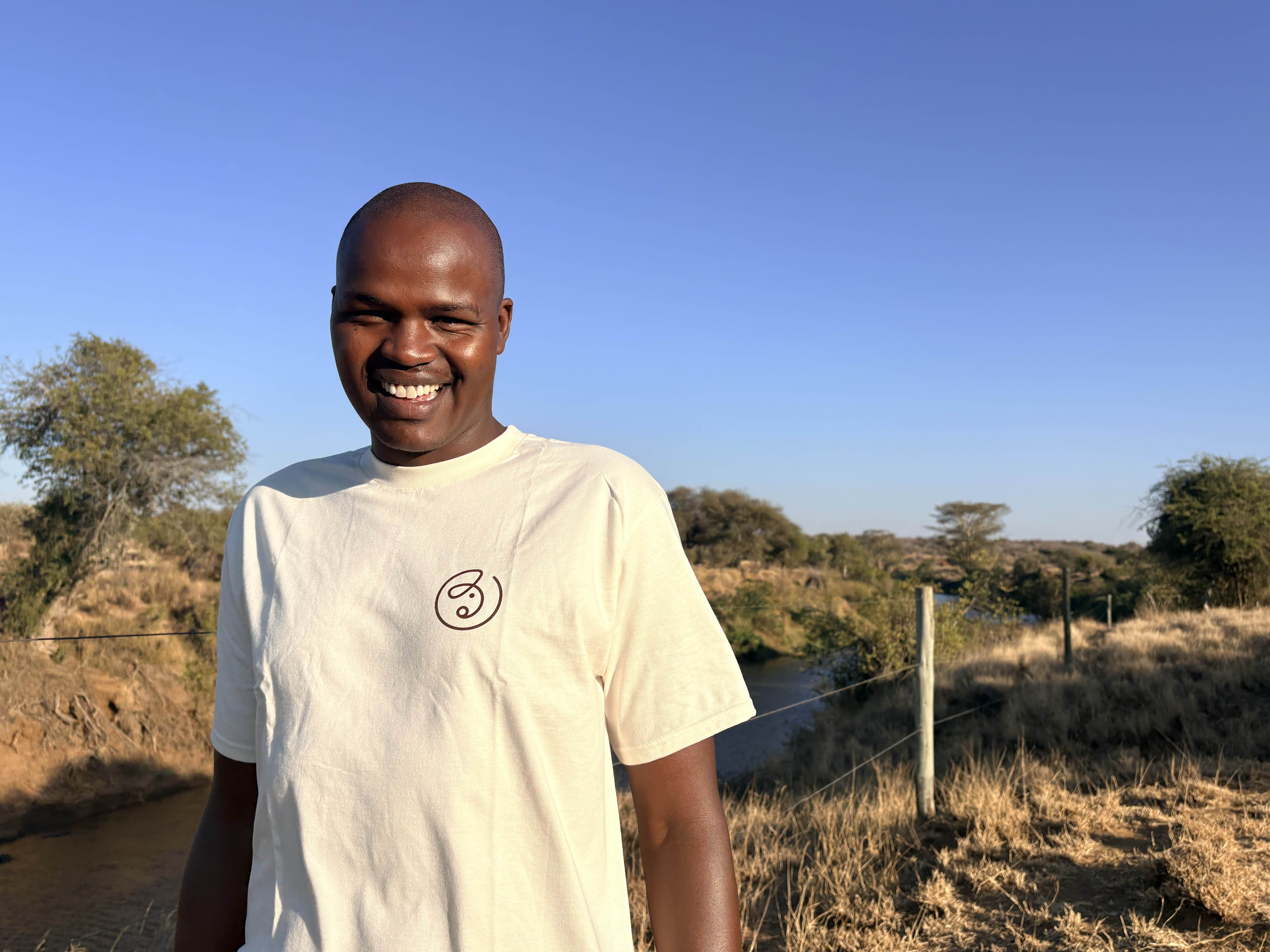 Smiling man wearing a light beige t-shirt standing outdoors in a dry grassy area with trees and a river under a clear blue sky.