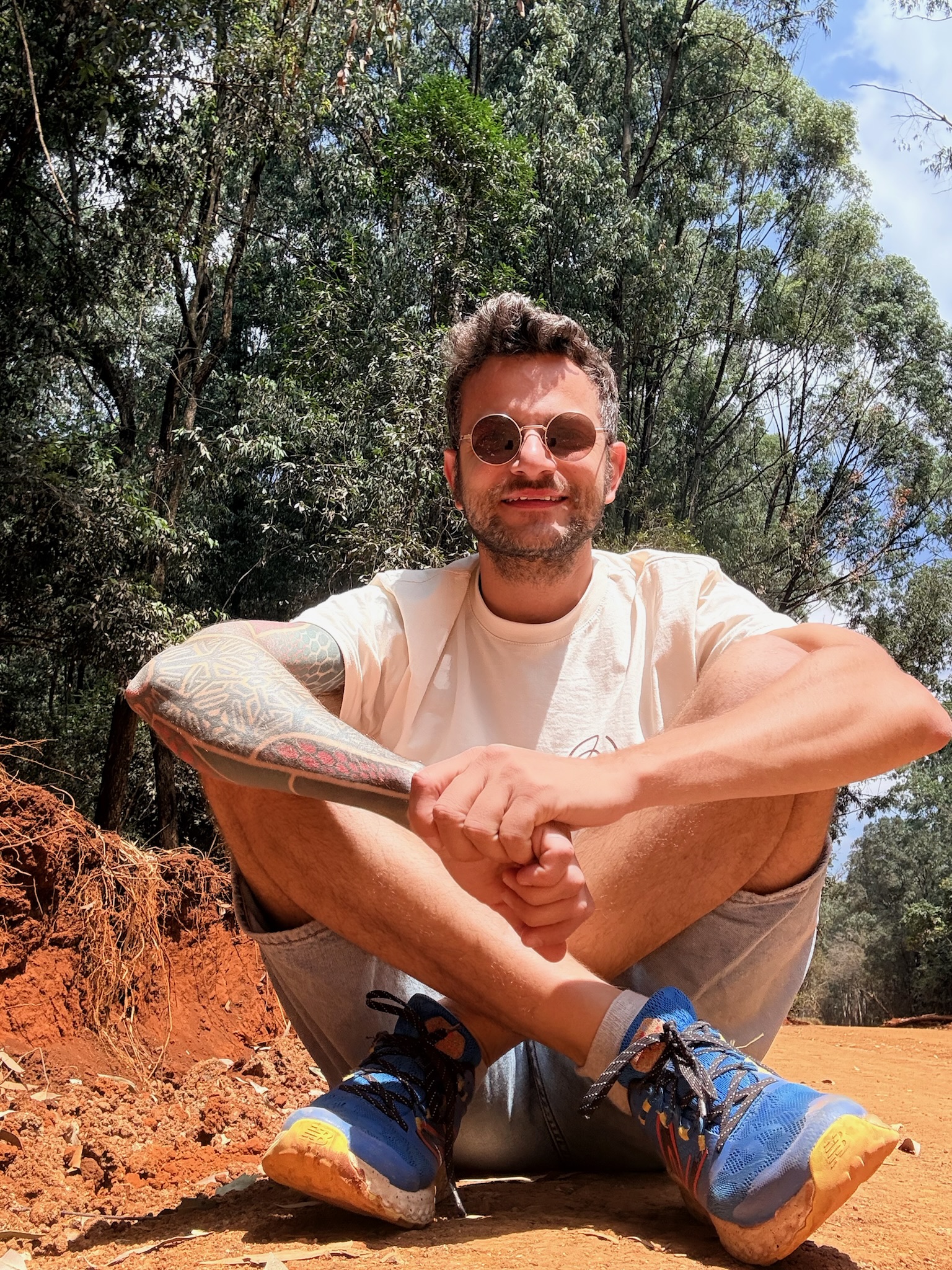 Man with round sunglasses, a light t-shirt, shorts, and blue sneakers sitting cross-legged on a dirt path surrounded by trees and under a partly cloudy sky.