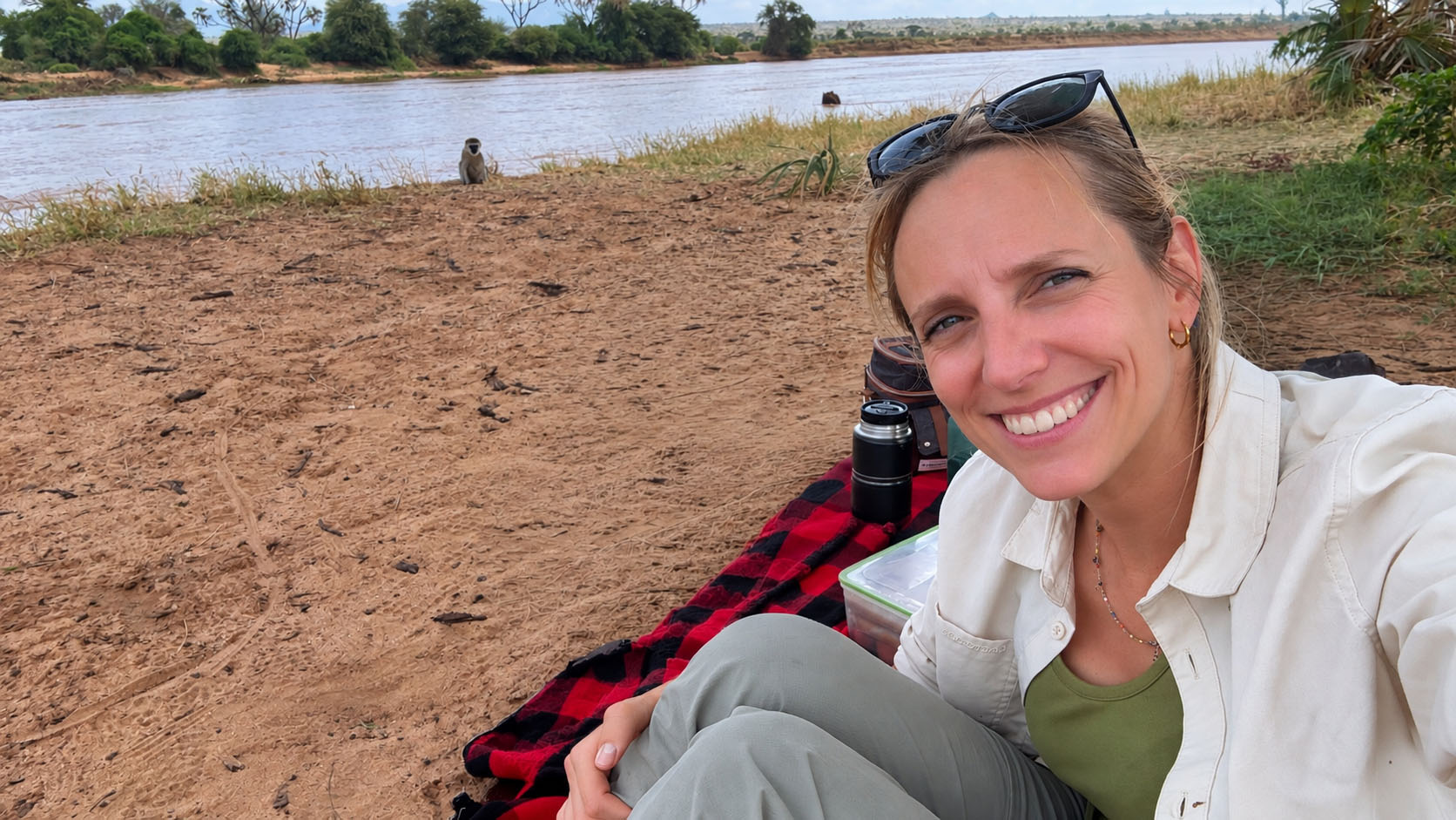Smiling woman with sunglasses on her head sitting on a red and black checkered blanket near a riverbank with sandy ground and green vegetation in the background.