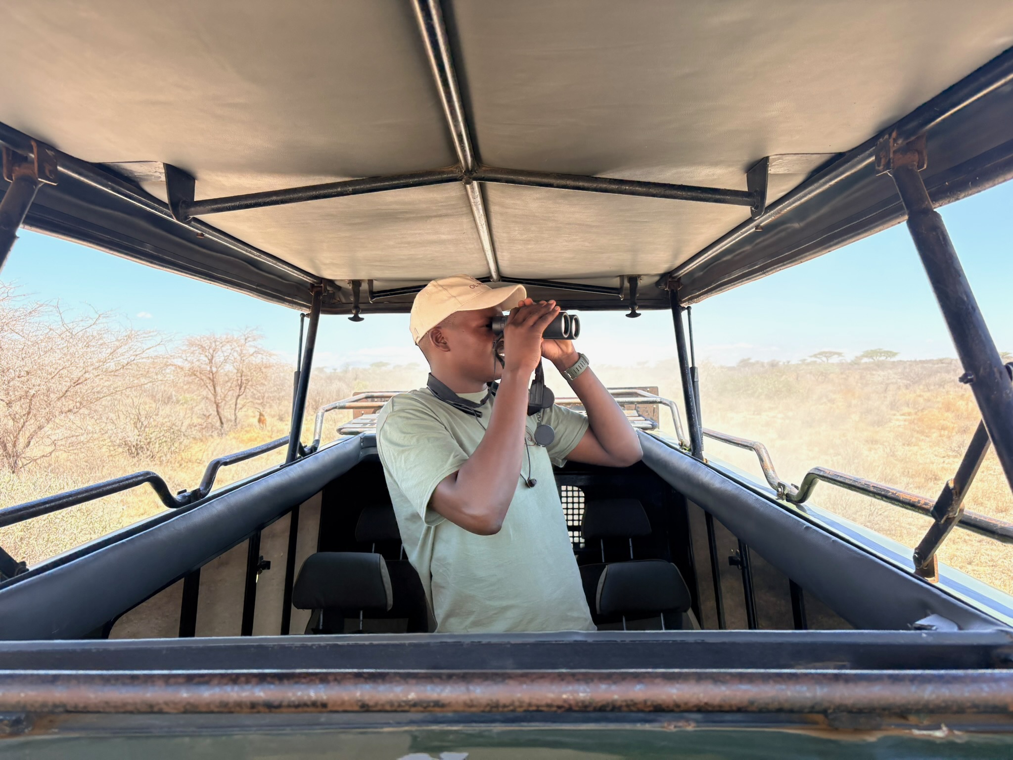 Man wearing a beige cap and green t-shirt standing inside an open safari vehicle, looking through binoculars at a dry, bushy landscape outside.