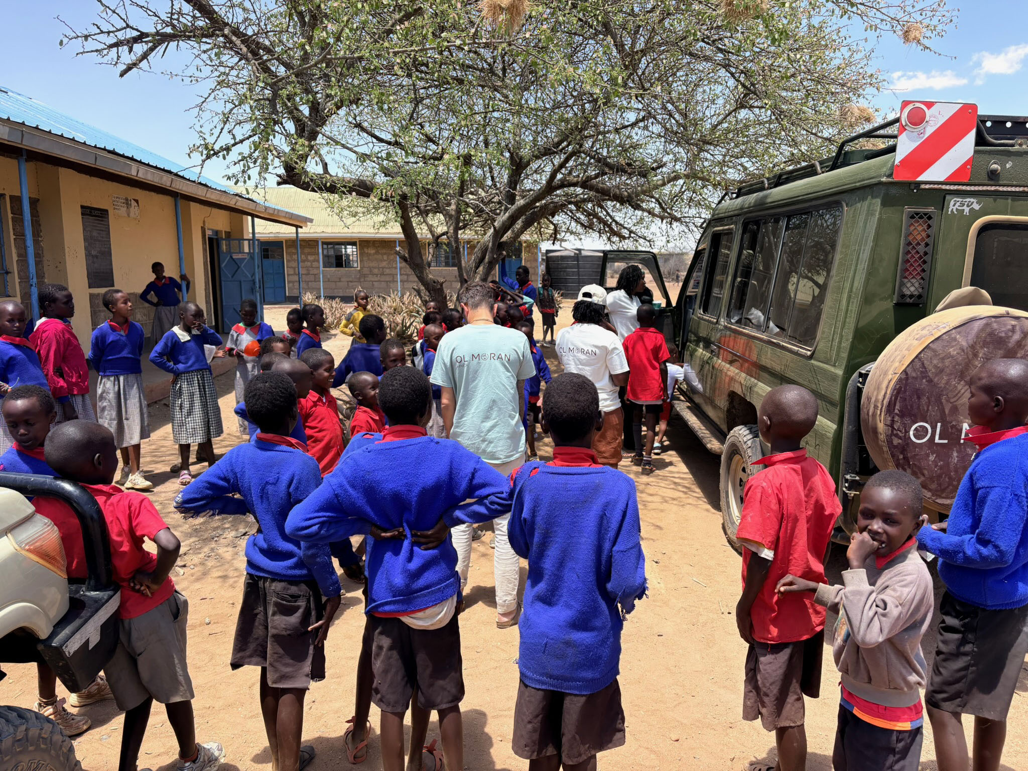 Group of school children wearing red shirts and blue sweaters standing outdoors around two adults in light blue and white shirts near a green safari vehicle under a tree beside school buildings.