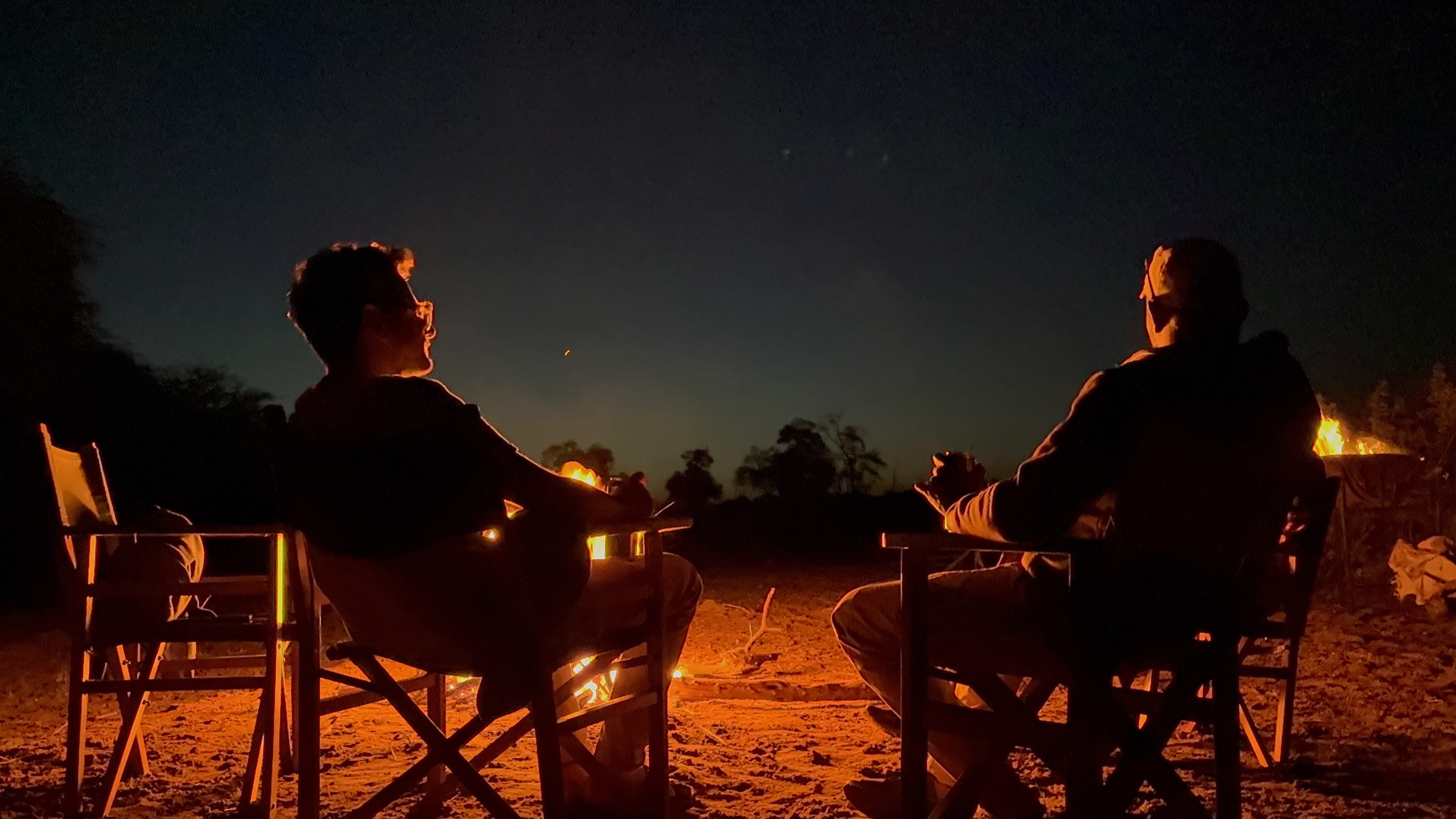 Two people sitting in chairs around a campfire at night, their silhouettes illuminated by the fire's warm orange glow against a dark sky with trees in the background.