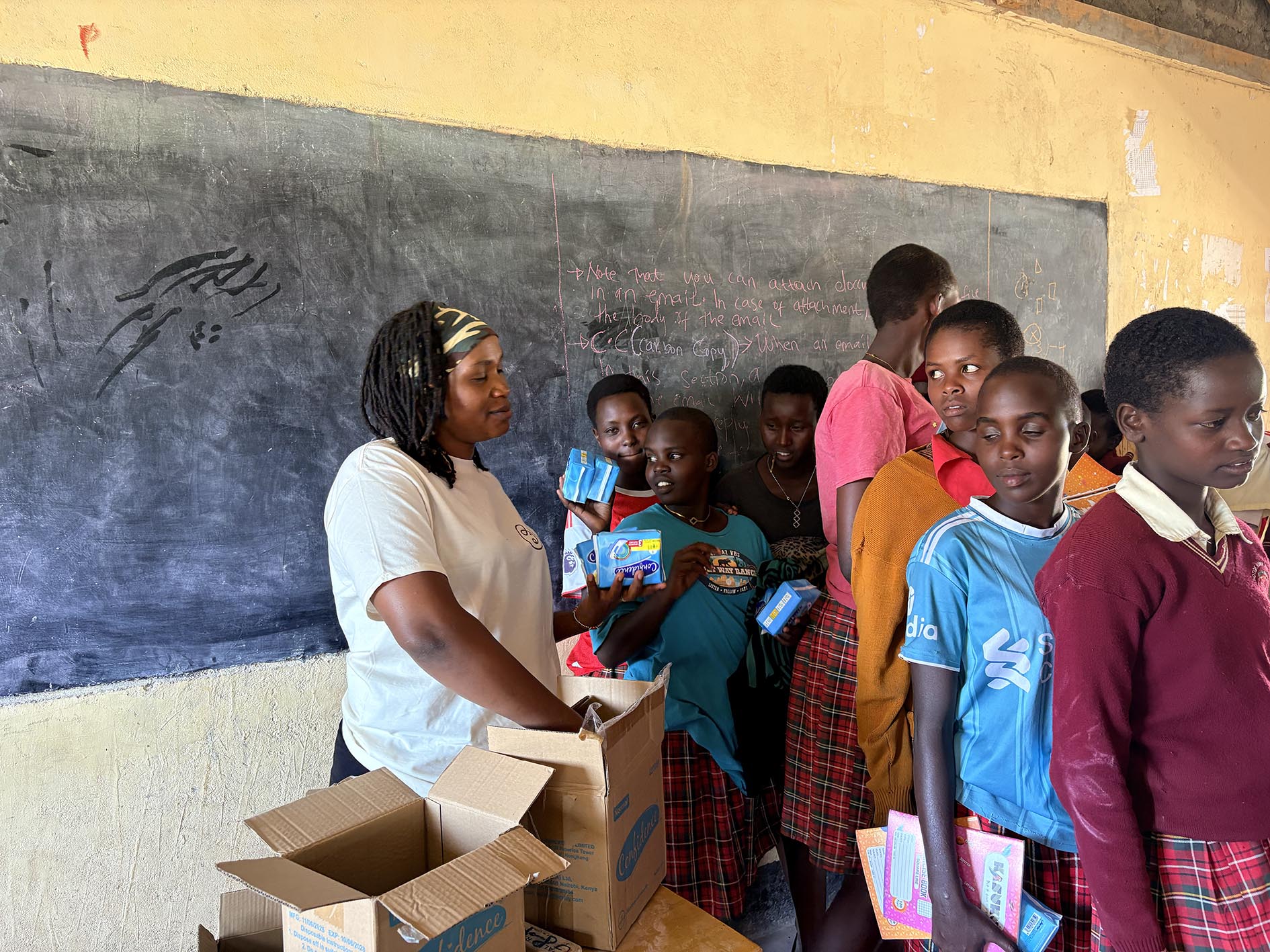 A woman in a white shirt distributes sanitary pad packs from open cardboard boxes to a line of girls dressed in school uniforms inside a classroom with a chalkboard in the background.