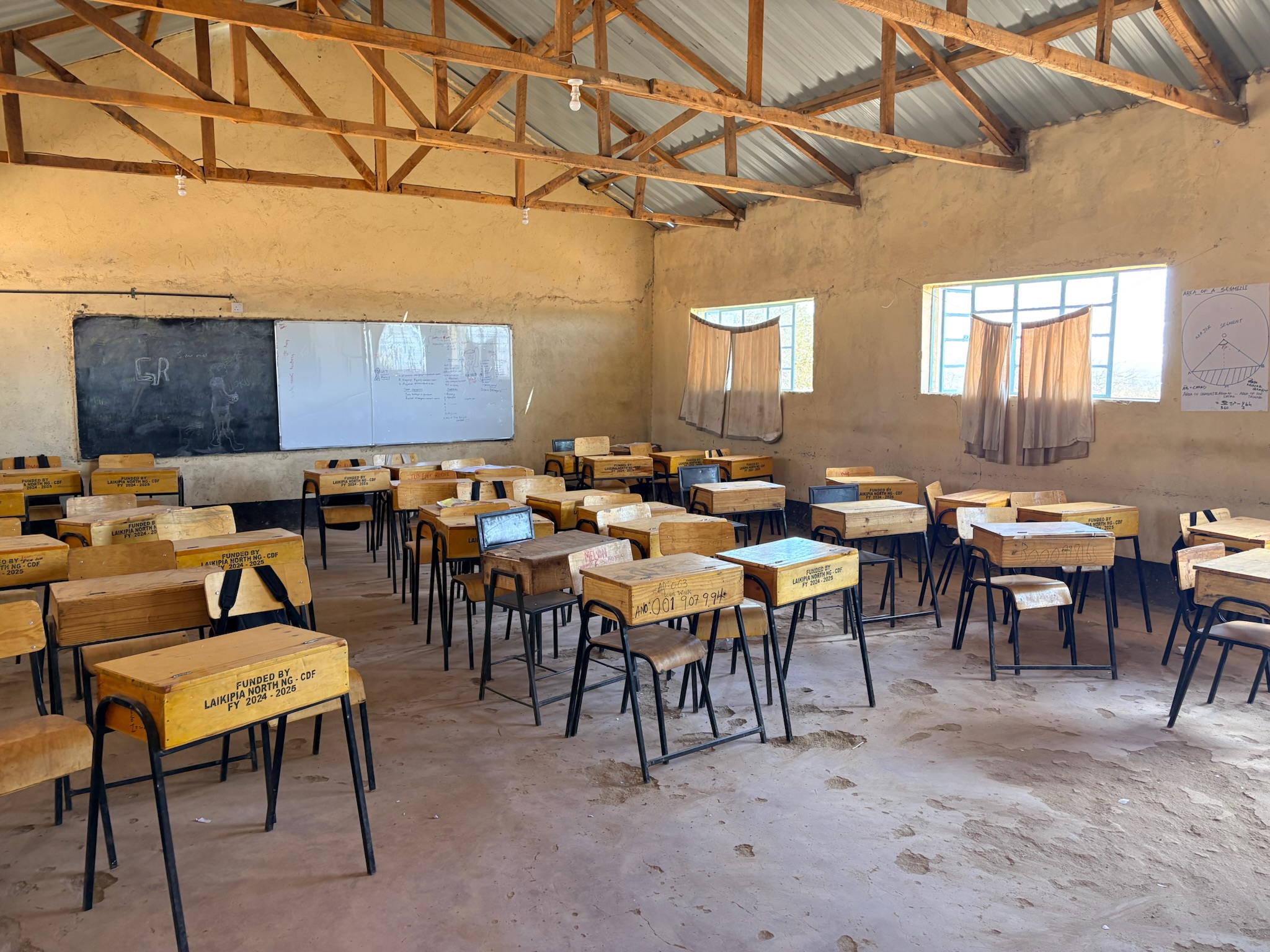 Empty classroom with wooden desks and chairs arranged in rows, two windows with beige curtains, blackboard and whiteboard on the front wall, and a metal roof with wooden trusses.