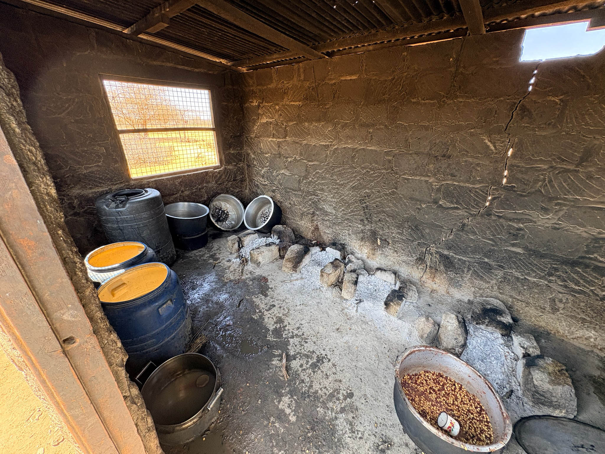 Interior of a rustic kitchen with a dirt floor, stone fire pit along one wall with ashes, several plastic and metal containers, a metal pot with beans and a cup inside it, and a window with a wire mesh.
