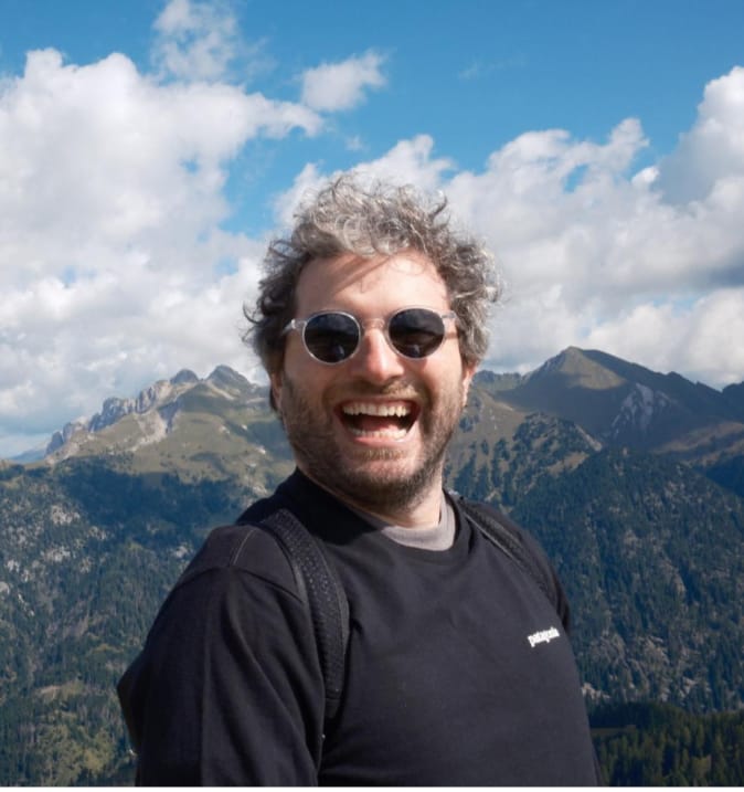 Smiling man with curly hair wearing round sunglasses and a black sweatshirt standing outdoors with mountains and partly cloudy sky in the background.