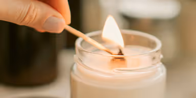 Hand lighting a white candle with a wooden matchstick in a glass container.