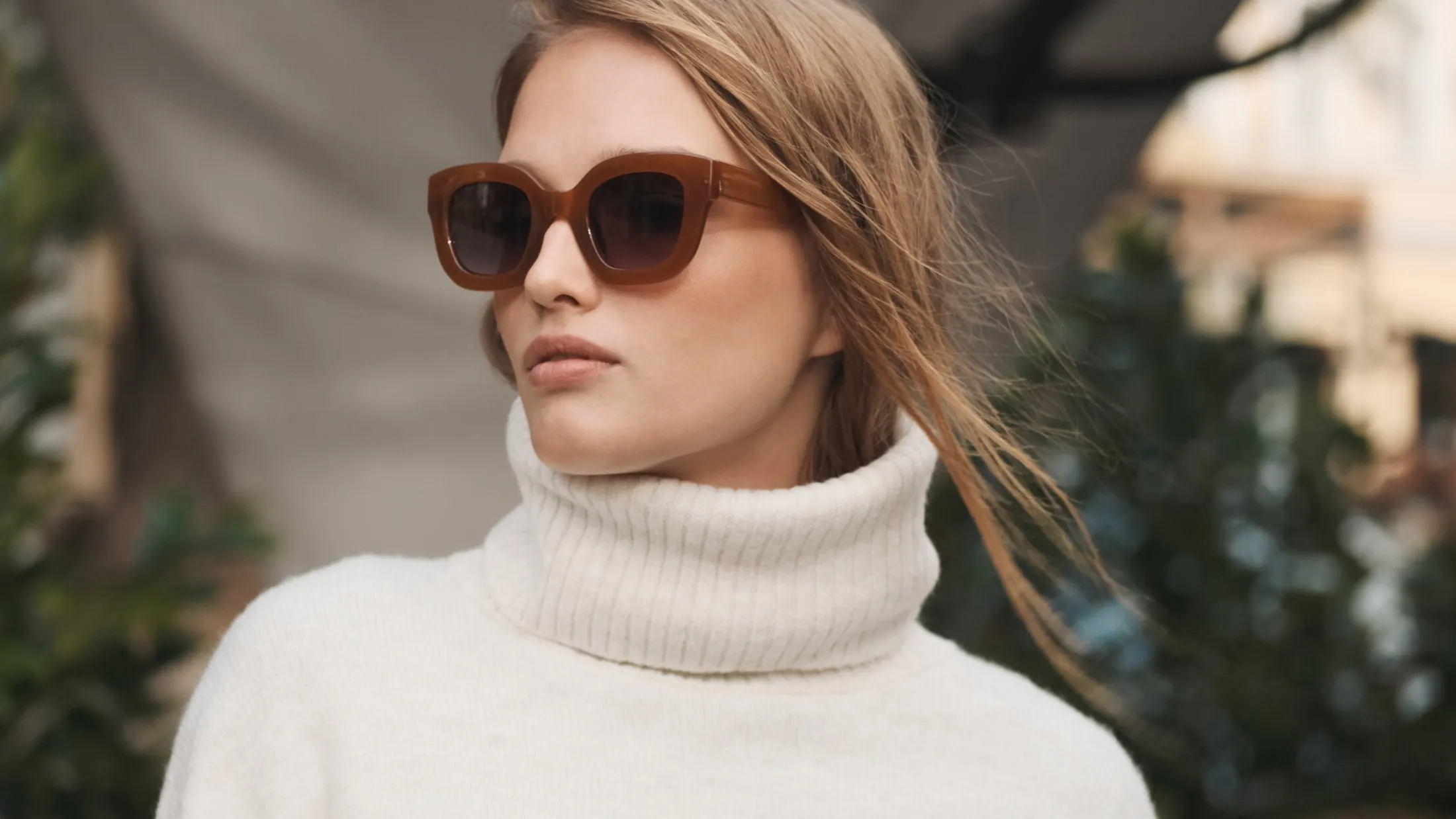 Young woman with light blonde hair wearing oversized brown sunglasses and a white ribbed turtleneck sweater, looking to the side with a neutral expression.