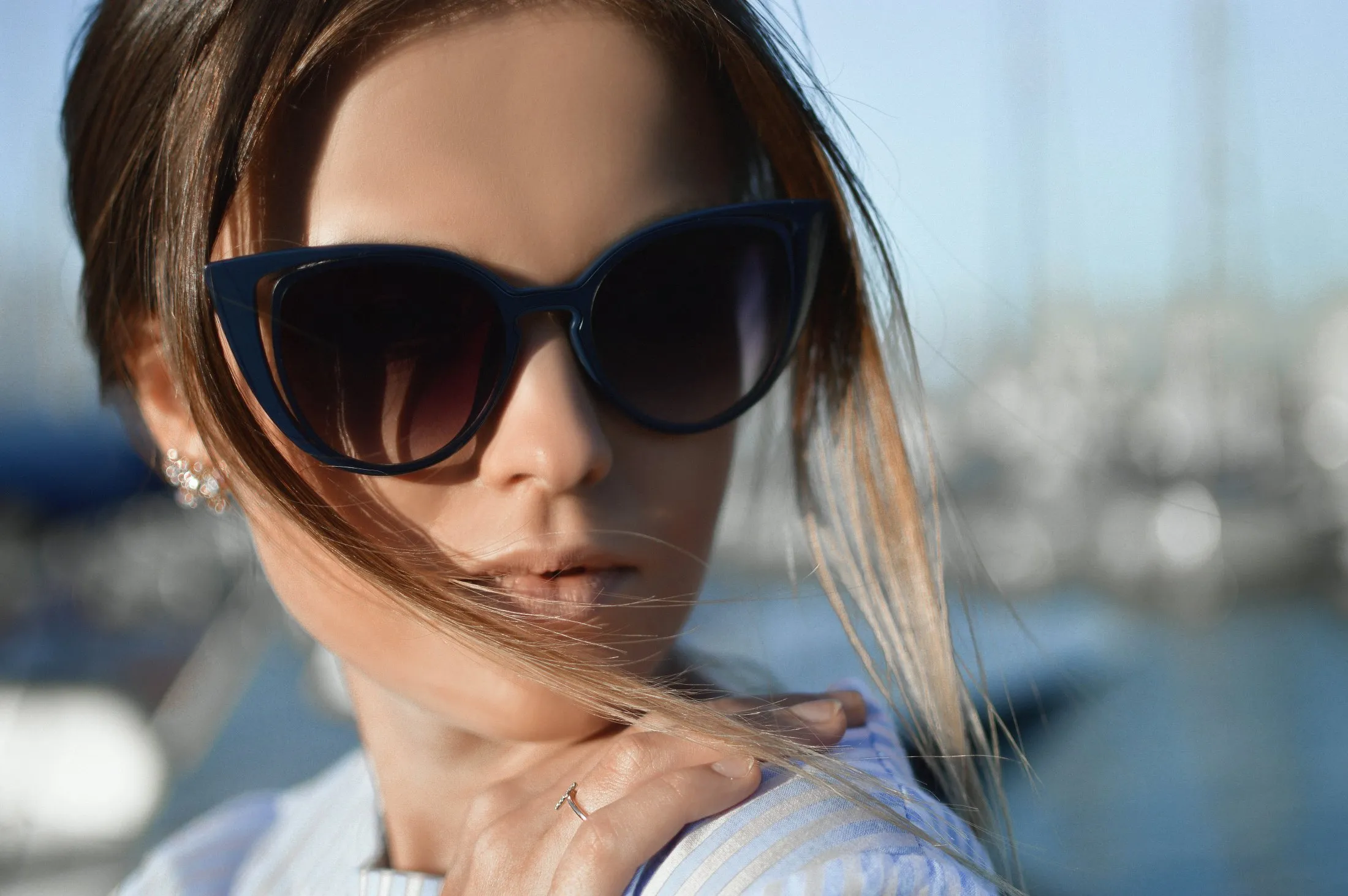 Close-up of a woman wearing large, dark sunglasses with light brown hair partially covering her face against a blurred outdoor background.