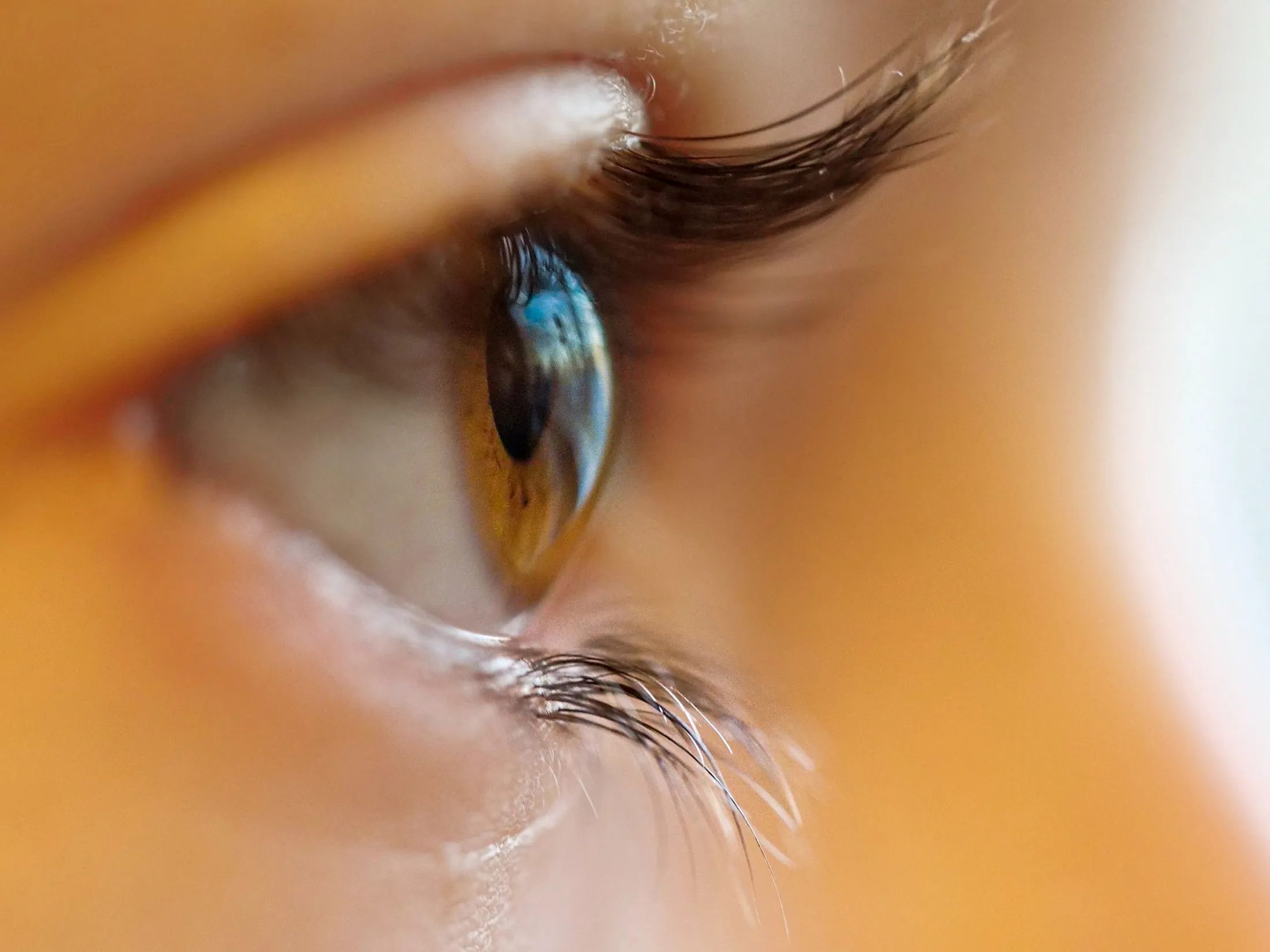 Close-up side view of a human eye with brown iris and long eyelashes, reflecting a window with blue sky and clouds.