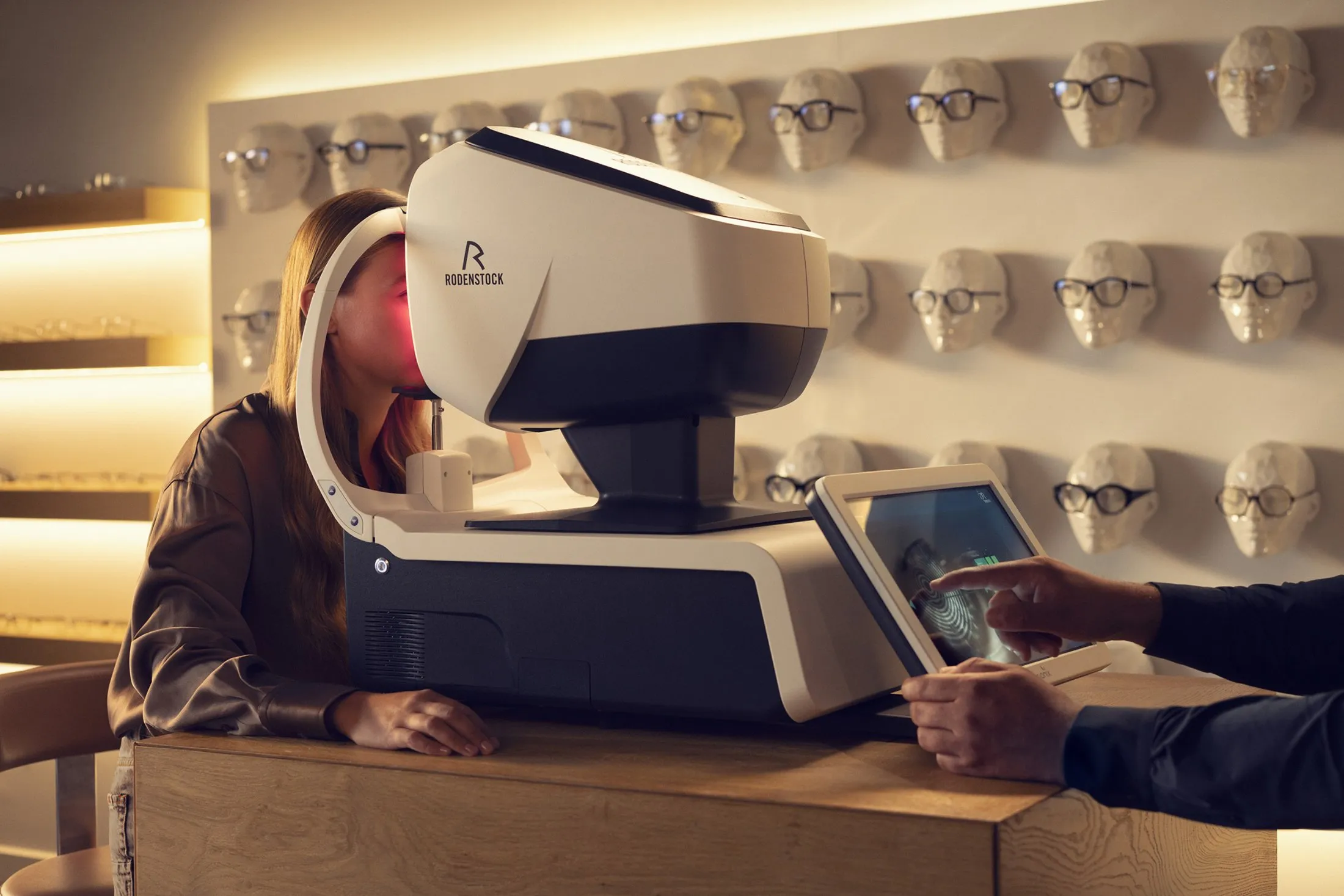 A woman with long hair uses a Rodenstock eye examination device while a technician operates the touchscreen interface, with mannequin heads wearing glasses displayed on the wall behind them.