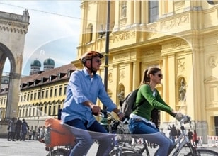 Man and woman riding bicycles side by side on a city street with historic buildings in the background.