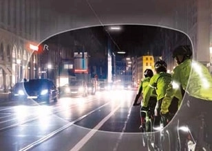 Nighttime view through a tinted lens showing a group of cyclists wearing reflective yellow jackets riding in a bike lane alongside a busy road with cars and trucks with headlights on.