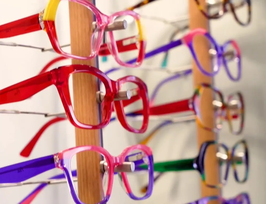 Colorful eyeglasses with transparent frames displayed on a wooden stand in rows, featuring shades of red, purple, yellow, and green.