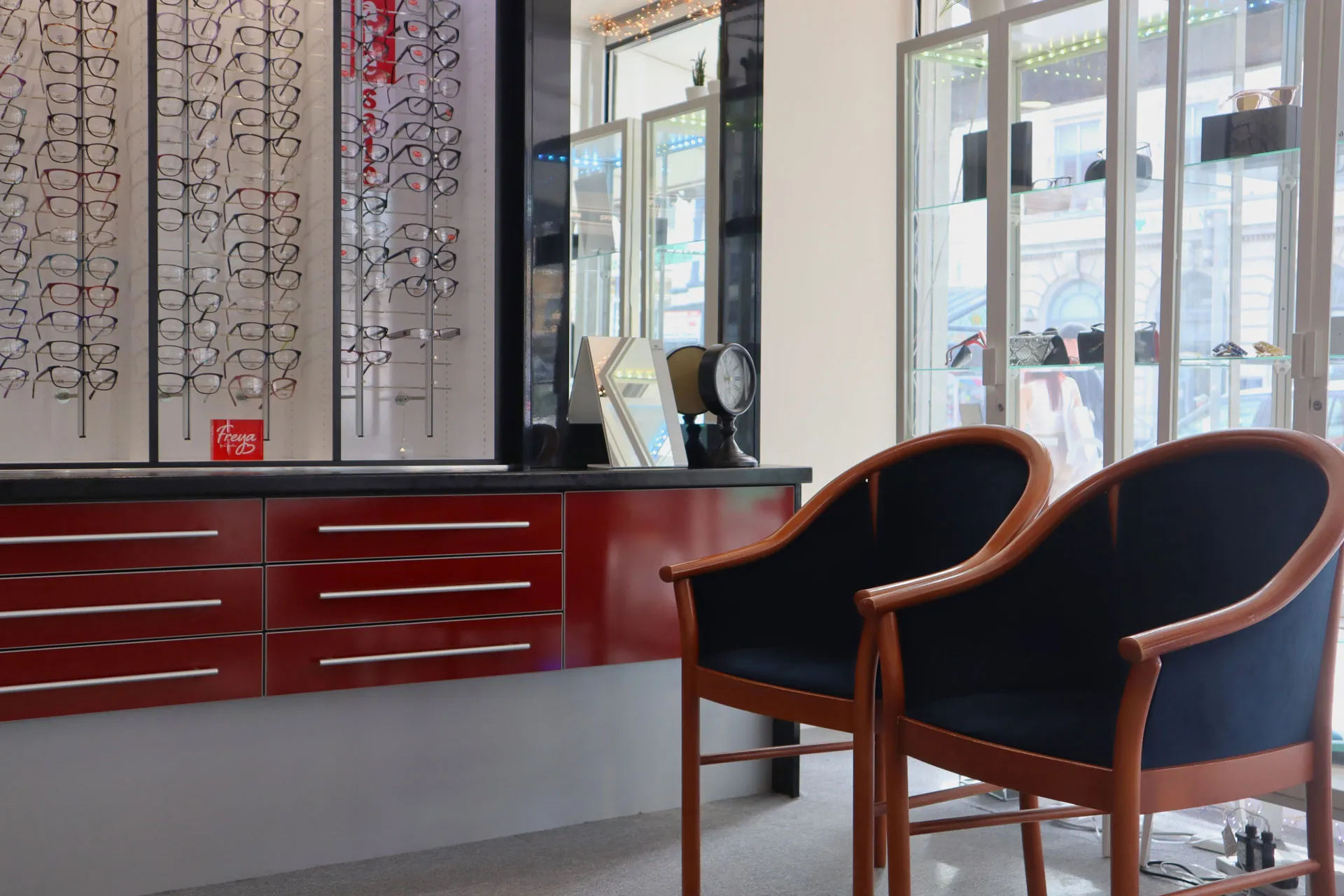 Interior of an eyewear store featuring red cabinets with metal handles, a display wall with various eyeglass frames, two wooden-framed chairs with dark cushions, and glass display cases near large windows.