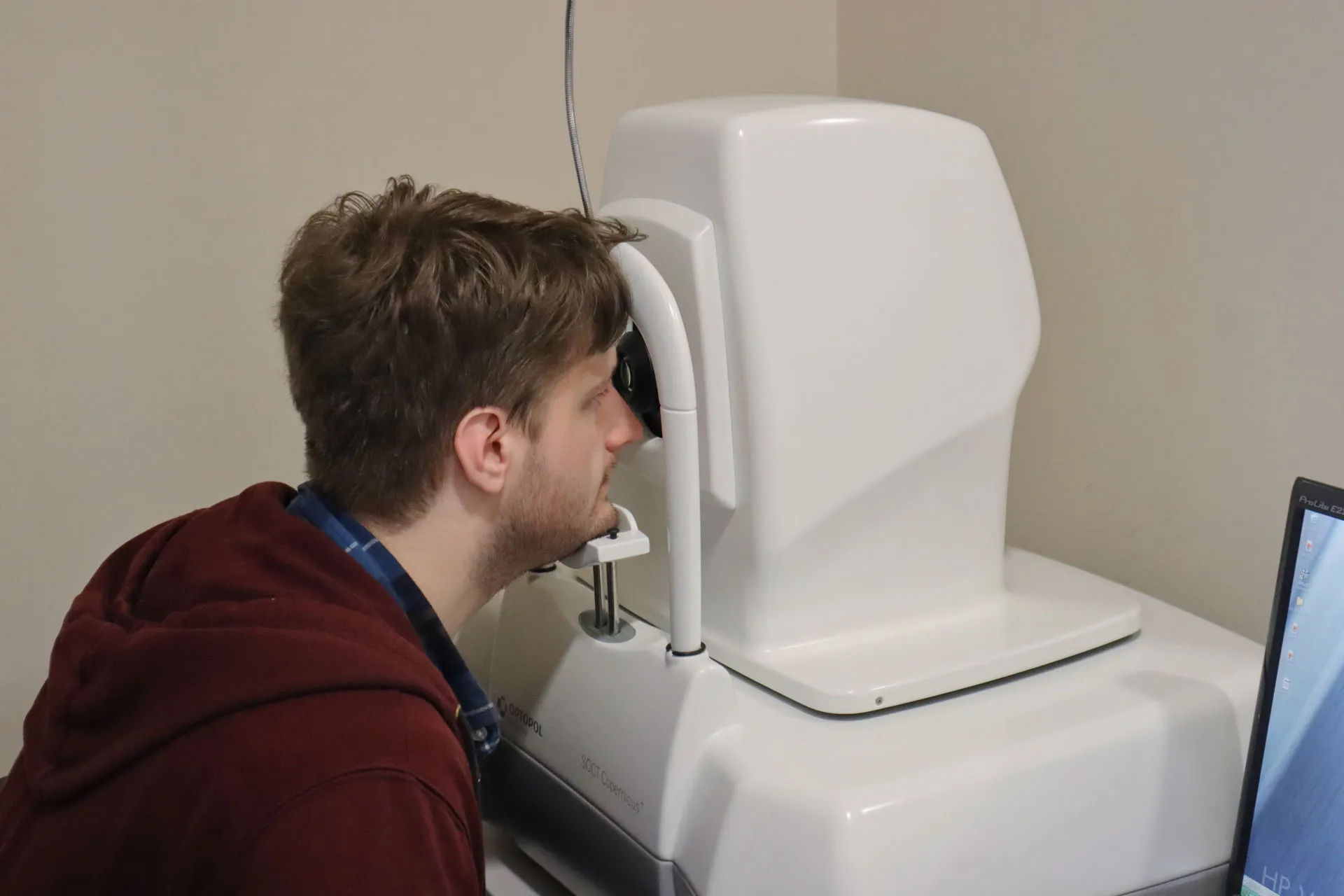 A man with short brown hair and a maroon hoodie rests his chin on a support while looking into a white ophthalmic device for an eye examination, with a computer monitor partially visible on the right.