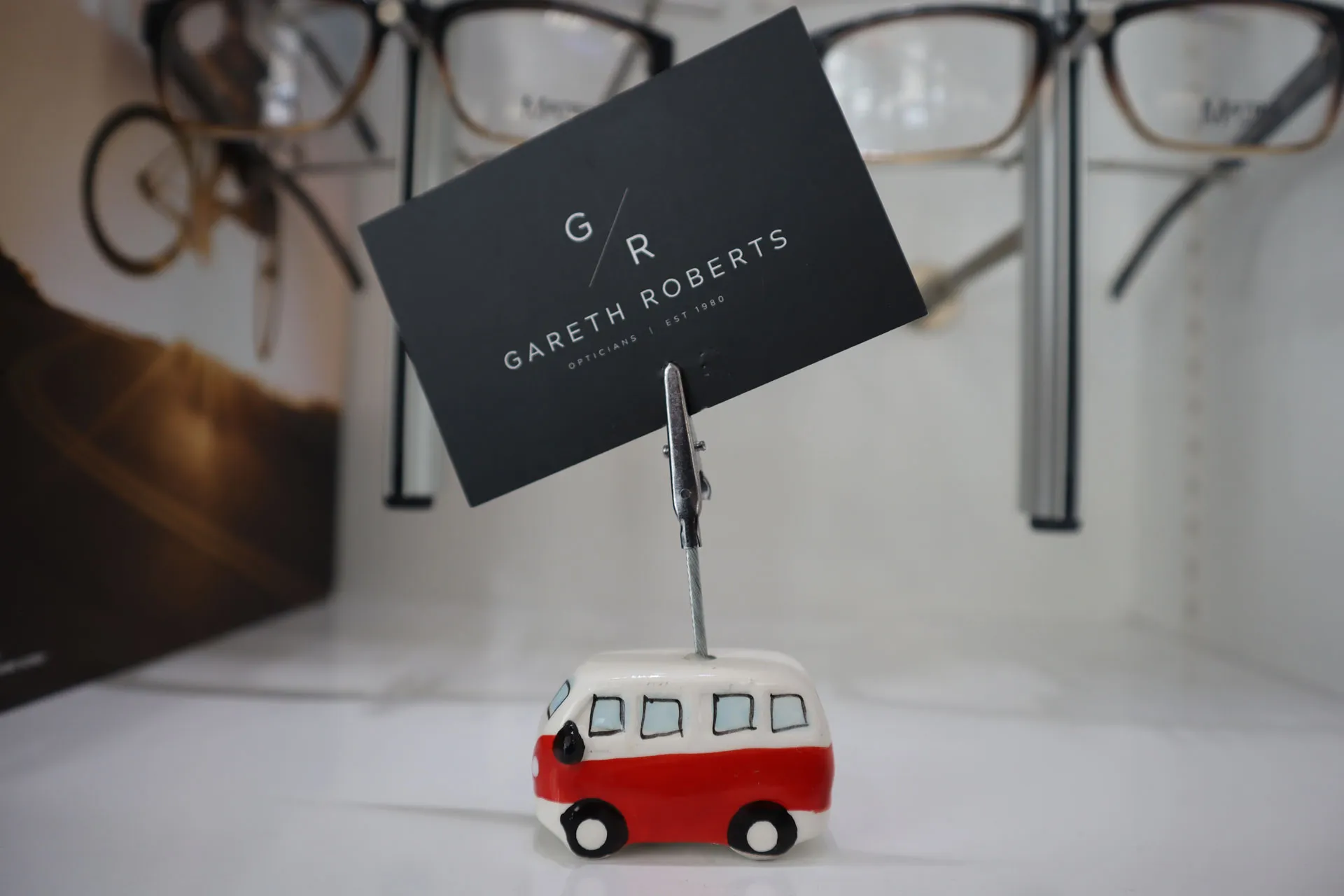 Small ceramic red and white van-shaped card holder holding a black card with 'Gareth Roberts Opticians EST 1980' text, set on a white surface with eyeglasses blurred in the background.