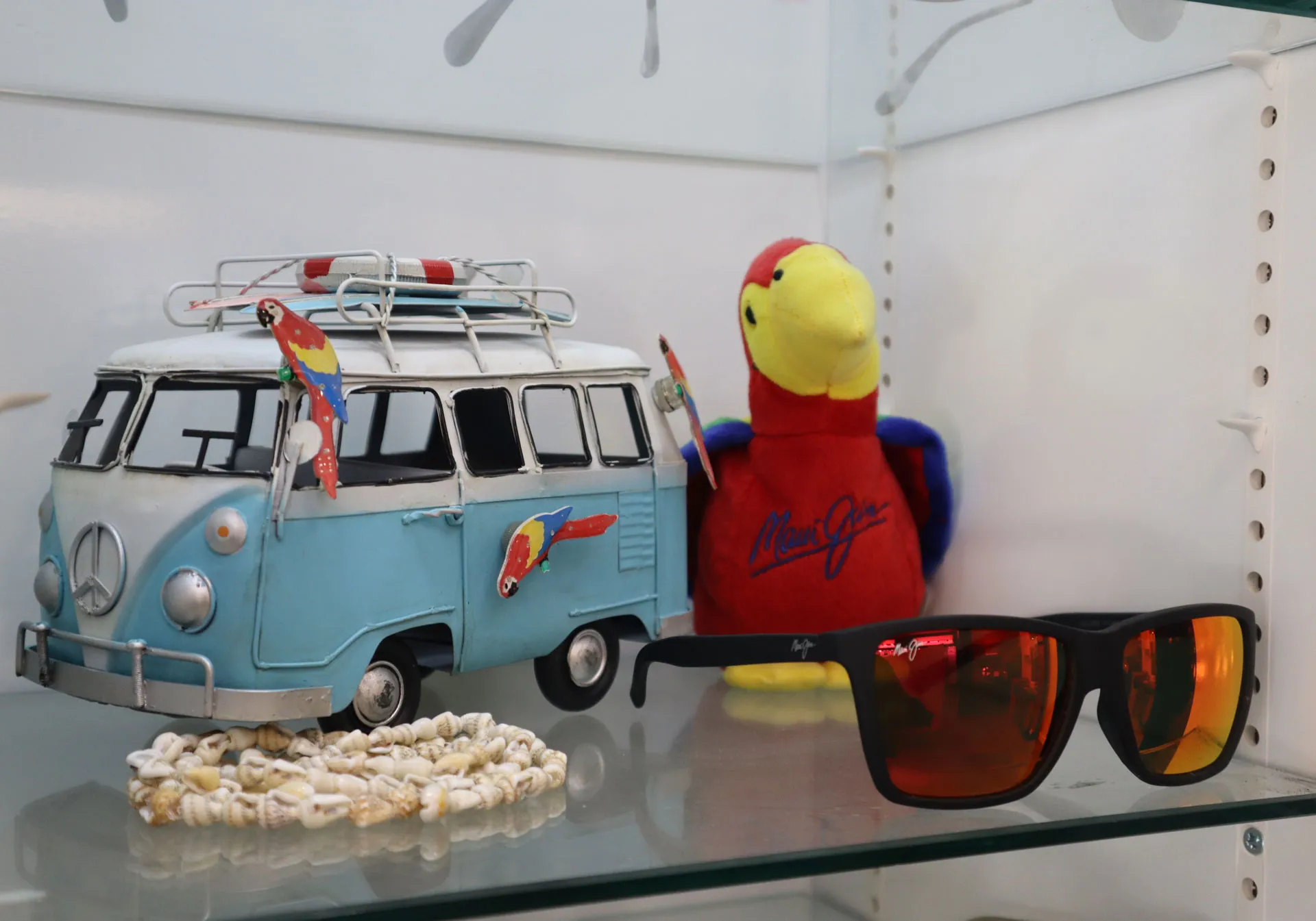 A light blue metal toy van decorated with colorful parrot figurines sits on a shelf next to a red and yellow plush parrot with 'Maui Jim' embroidered in blue, and a pair of black sunglasses with orange reflective lenses.