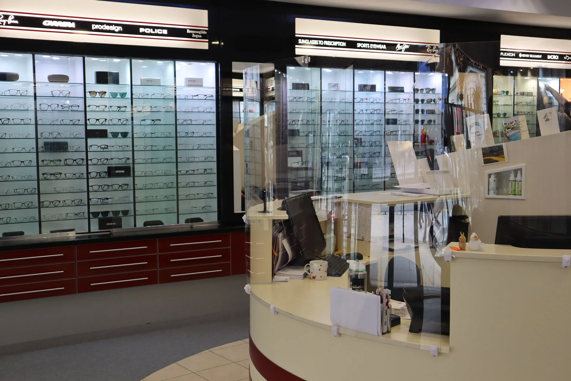 Interior of an eyewear store with multiple glass display cases filled with various eyeglasses and sunglasses, a curved reception desk with a transparent protective screen, computer monitors, a keyboard, a mug, office supplies, and brochures.