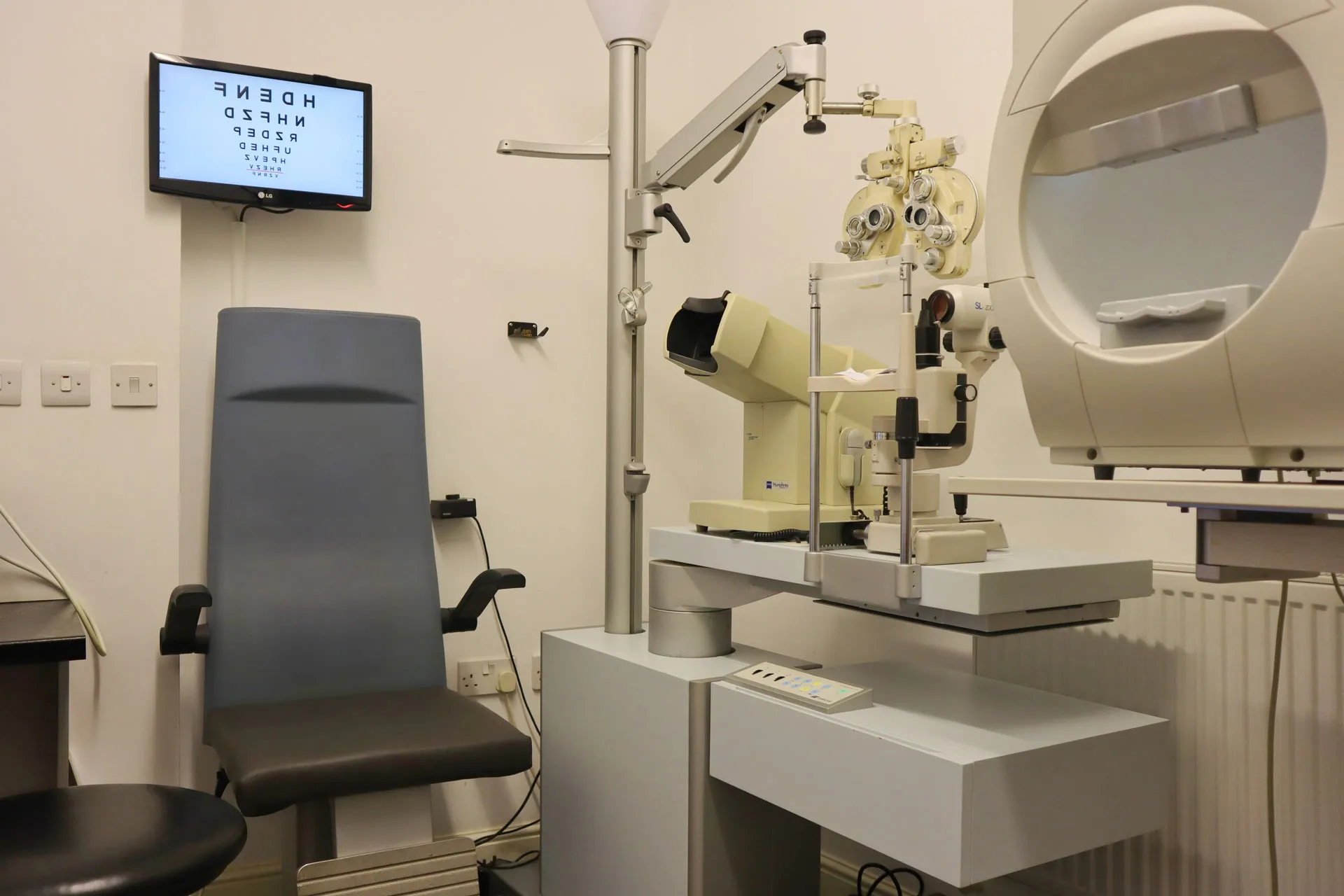 An optometrist exam room featuring a patient chair, an eye chart on a wall-mounted screen, and ophthalmic diagnostic equipment including a phoropter and slit lamp.