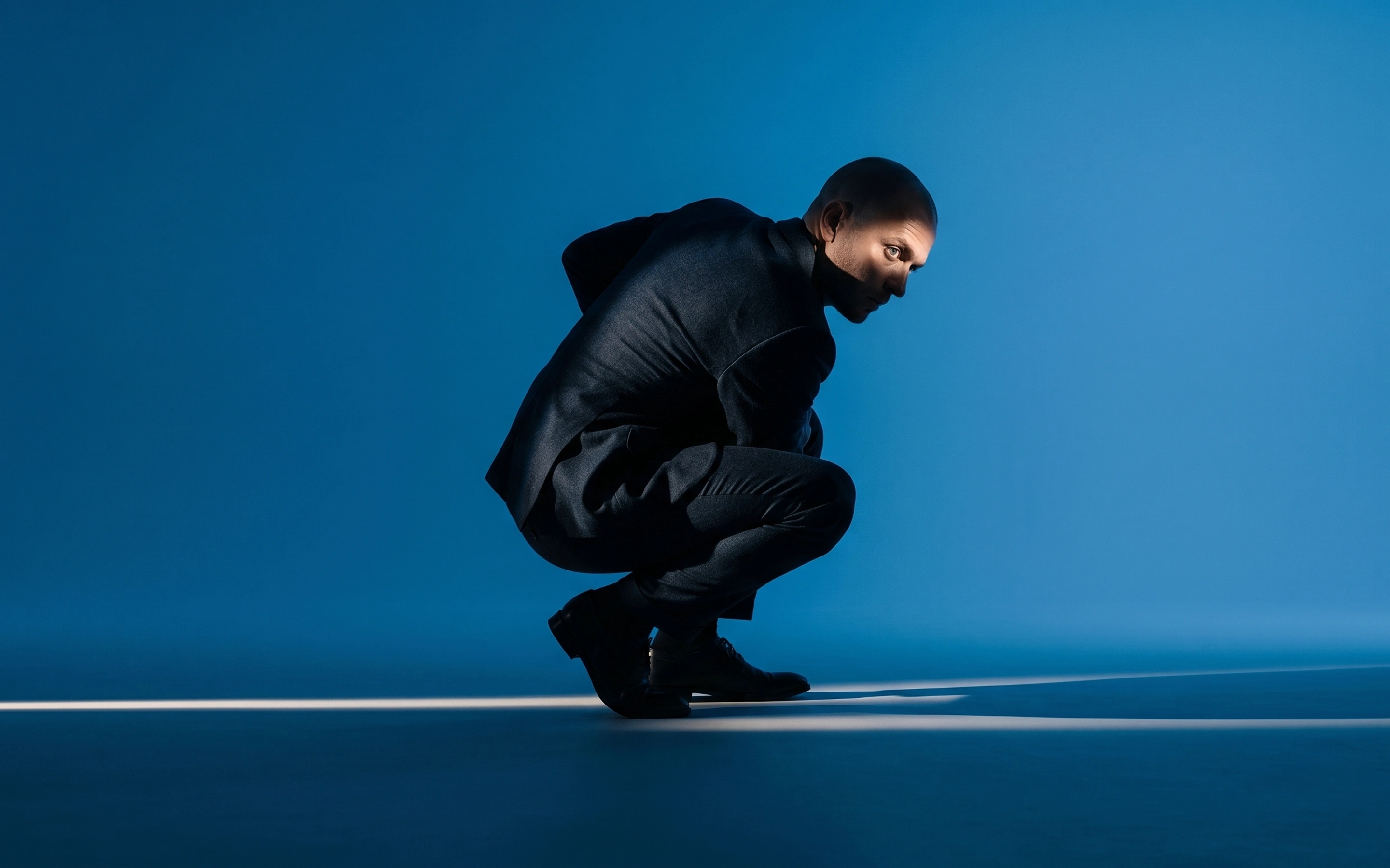 Man in a dark suit crouching on a blue floor with a focused expression in spotlight.