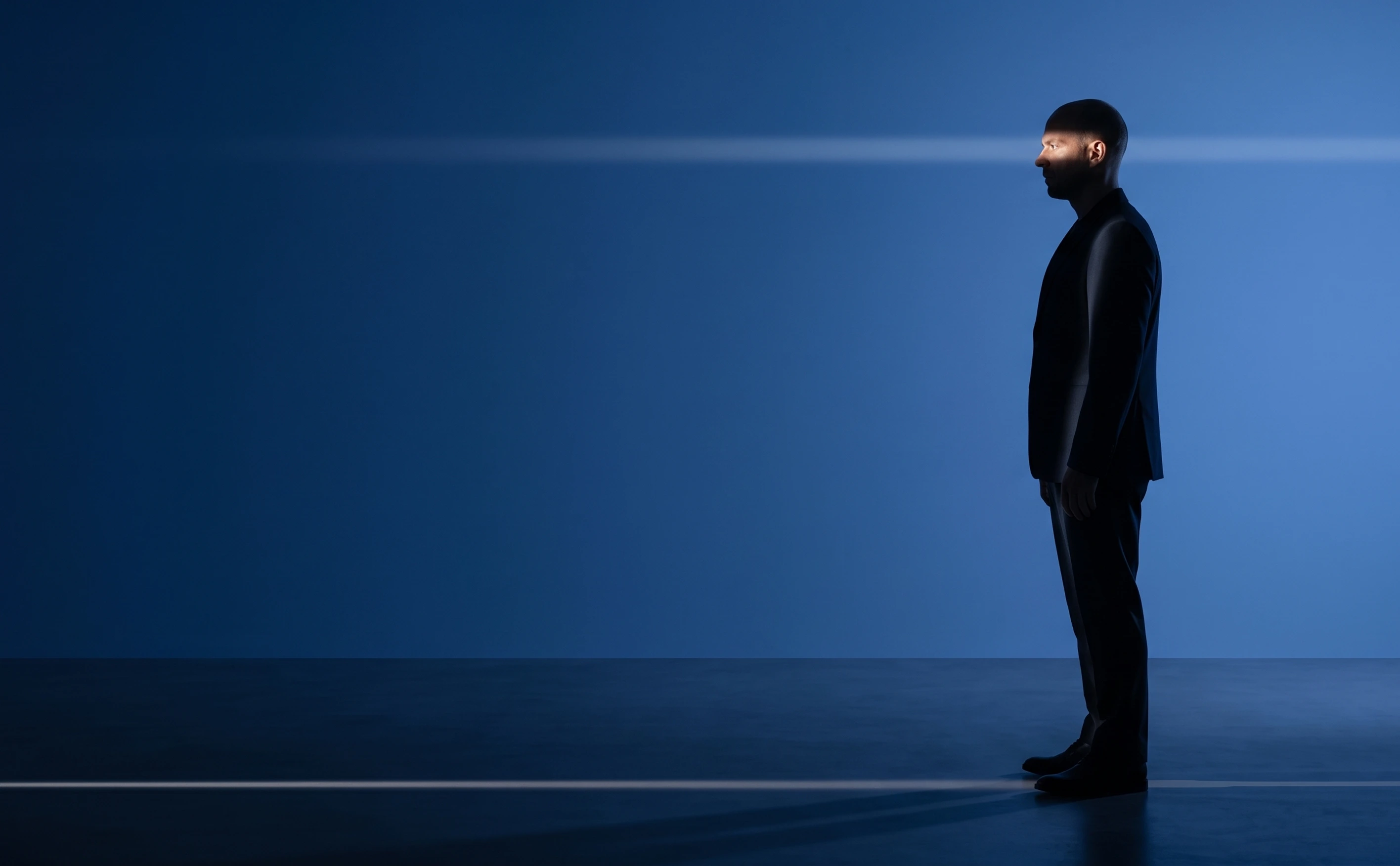 Man in black suit standing sideways with a horizontal beam of light crossing his face and eyes.