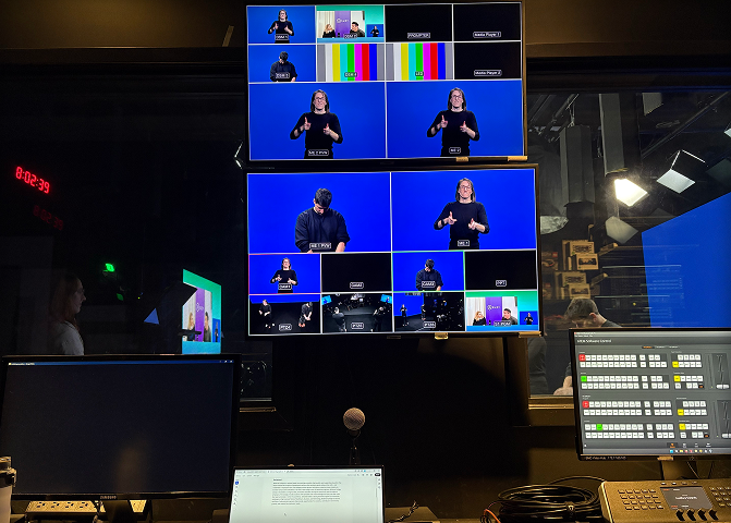 Broadcast control room with screens showing sign language interpreters and a control panel with buttons and a microphone.