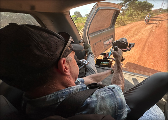 A man inside a vehicle capturing video of a motorcyclist on a dirt road using a camera rig.
