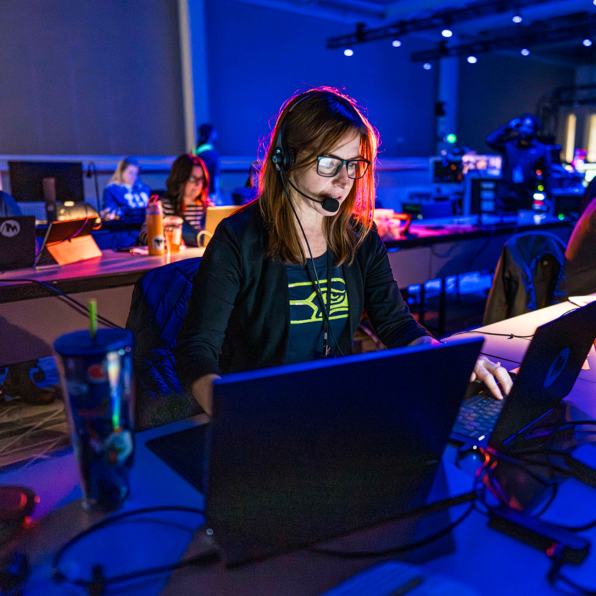 Woman wearing headset and glasses working on a laptop in a dimly lit tech workspace.