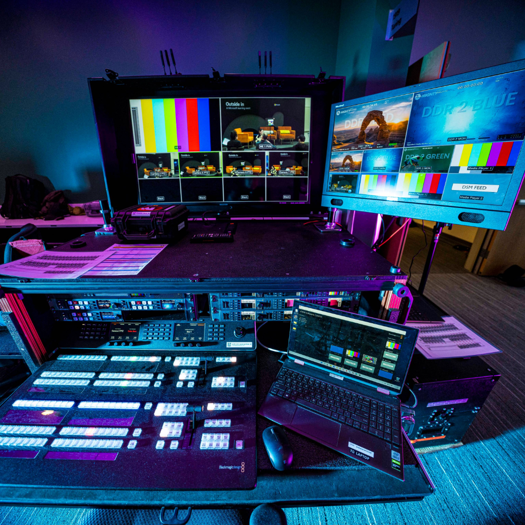 Video production control station with monitors, switcher, laptop, and control panels lit in blue and purple.